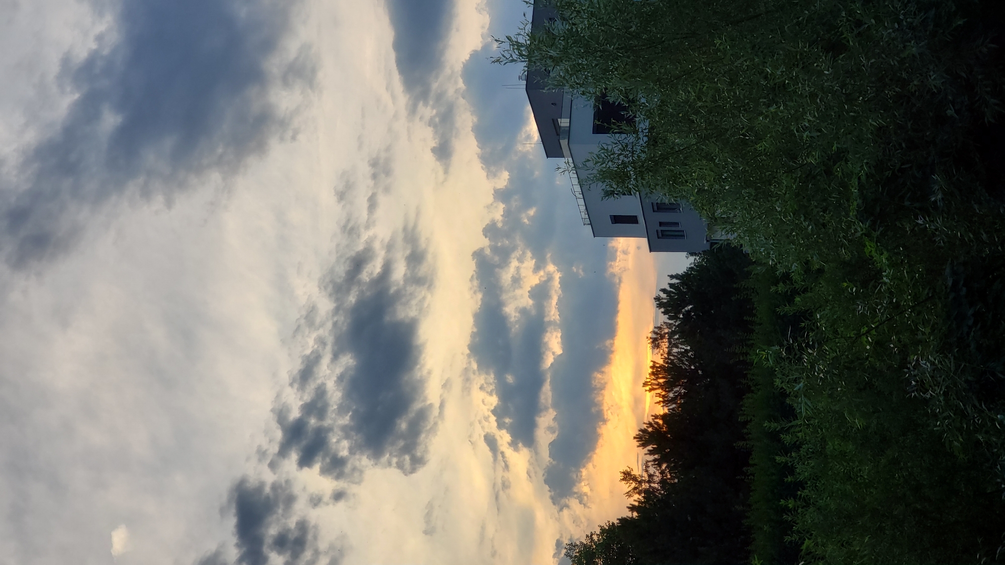 The image captures a twilight scene with a dramatic sky filled with clouds, tinged with the warm hues of sunset. A modern building, characterized by its white and gray facade with multiple windows, is visible on the right side of the frame, partially obscured by the lush green foliage of trees and bushes in the foreground. The trees in the middle ground form a dark silhouette against the brighter sky, with the setting sun's light filtering through their branches, creating a warm orange glow. The location is identified as the city of Prešov, Slovakia. The scene is static and devoid of any people or overt actions.