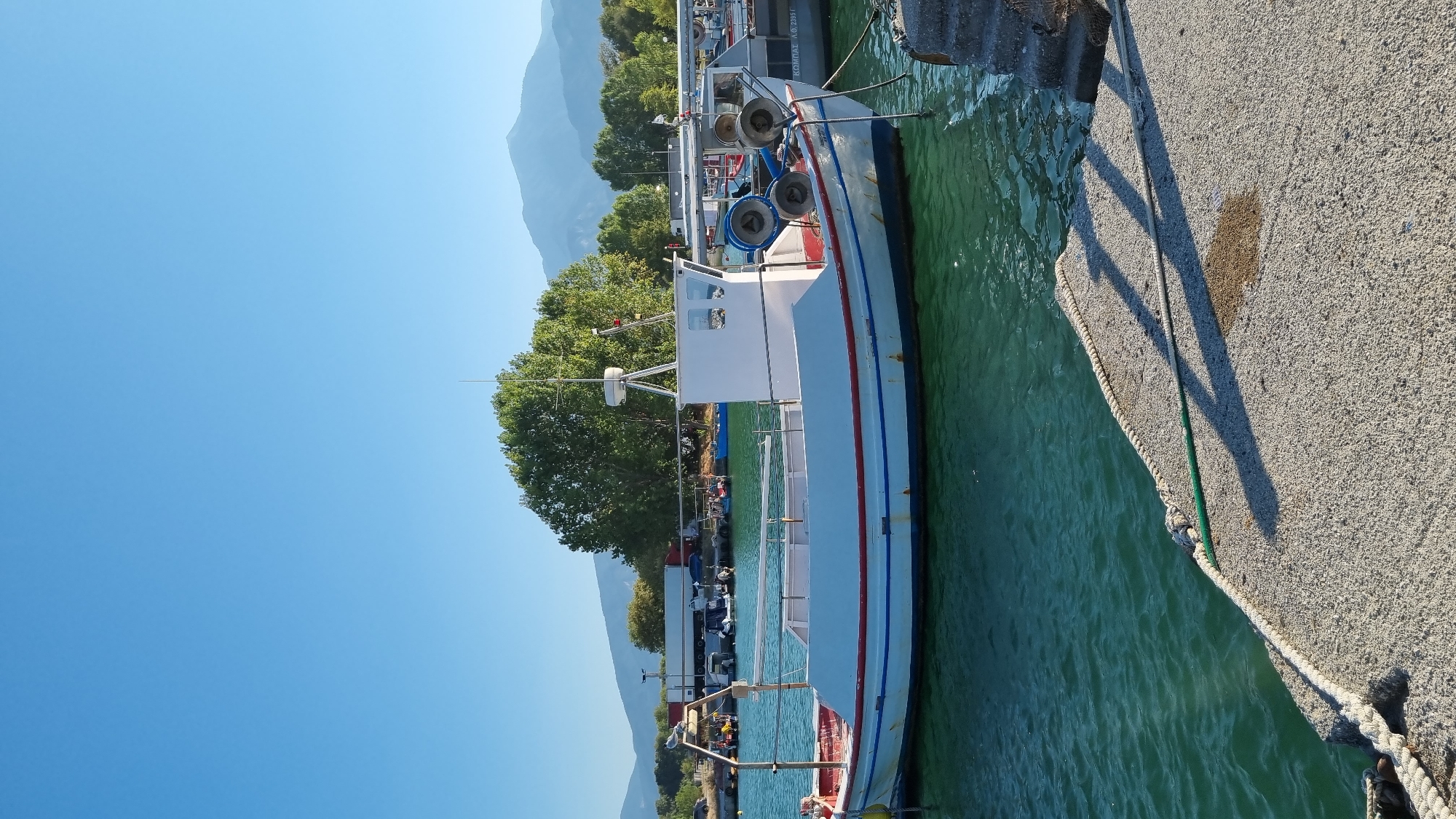 A clear, sunny day at the harbor of Limani Litochorou, Greece. A white fishing boat with red and blue trim is moored in the foreground. The boat has a cabin structure and several coiled ropes and buoys on its deck. The water is a vibrant green and appears calm. To the right, a concrete jetty with a rope extending from the boat is visible, along with some stone steps leading up. In the background, more boats are docked, and a line of trees stands against a backdrop of distant mountains under a clear blue sky. The overall scene depicts a peaceful maritime setting.
