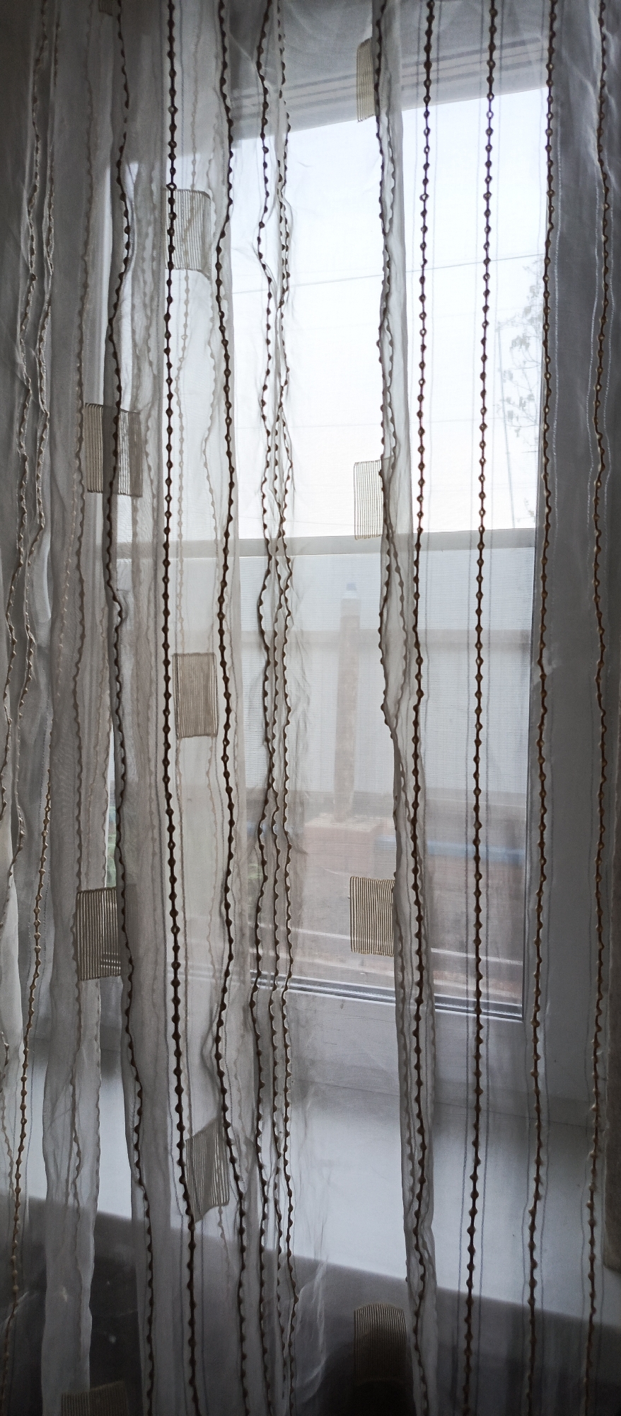 The image shows a close-up view of sheer, white curtains with vertical brown beaded trim and square decorative patches that appear to be woven with horizontal lines. The curtains are drawn across a window, allowing diffused light to enter. Through the translucent fabric, the outline of a window frame is visible, and beyond that, a blurry view of what appears to be an outdoor scene with some foliage and possibly a building or fence. The lower part of the image shows the windowsill and a hint of what might be the floor or ground outside. The style of the curtains and the view through the window suggest an interior domestic setting. Given the prompt's request for location specificity in Kalachinsk, Russia, this image likely depicts a window in a residential or public building within that city. However, no specific landmarks or identifying features within the image itself confirm the location. The overall mood is one of quiet domesticity or privacy.