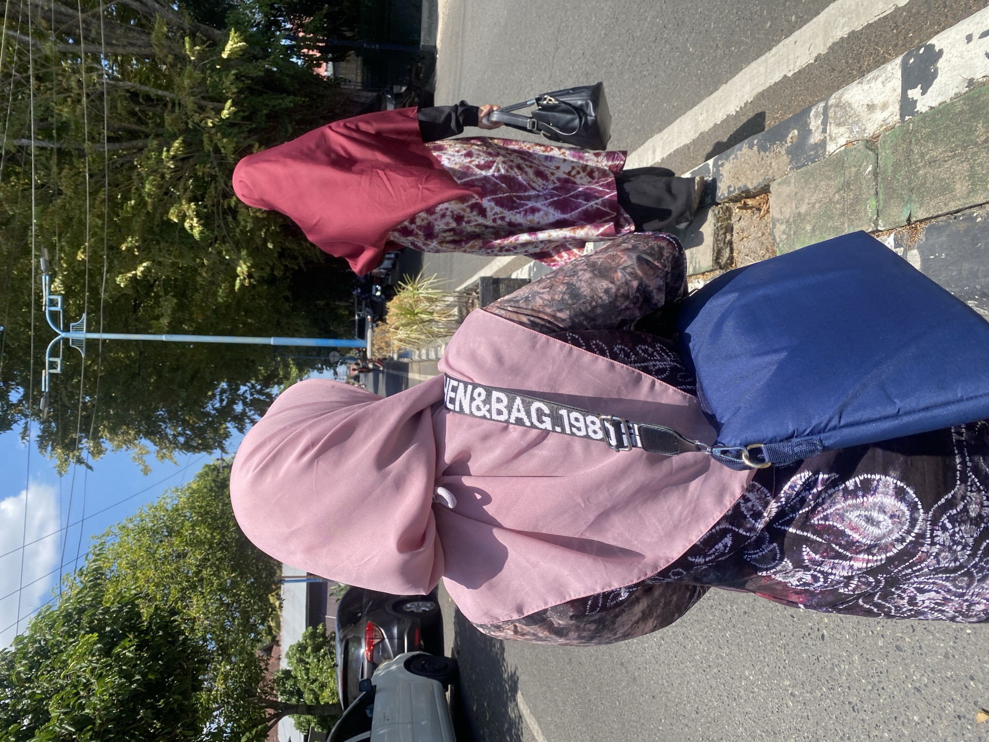 In Banjarbaru, Indonesia, two women walk along a street. The woman in the foreground wears a pink hijab and a patterned dress. A blue bag hangs from her shoulder, and a strap with "VEN&BAG.1981" is visible. The second woman, further down the street, wears a dark red hijab and a patterned dress over black pants. She carries a black handbag. Cars are parked along the street, and trees line the sidewalk. The scene is illuminated by sunlight under a bright blue sky with some clouds.