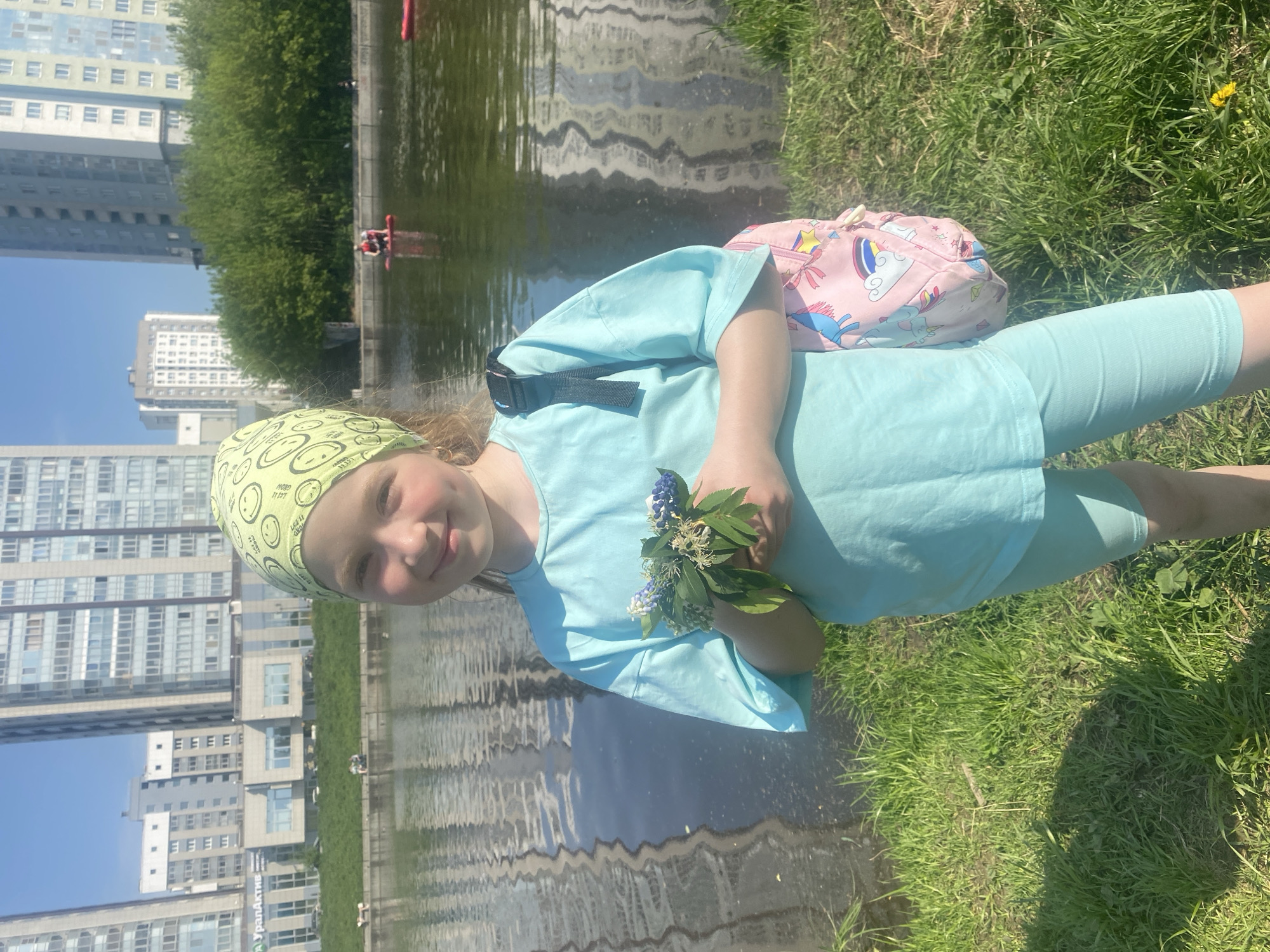 The image depicts a young girl standing beside a body of water, likely a pond or canal, in what appears to be an urban park setting. She is wearing a light blue t-shirt and matching leggings, with a lime-green bandana with a smiley face design. She is holding a small bouquet of wildflowers in her hands. A pink backpack with cartoon unicorn designs rests on the grass near her. In the background, a body of water reflects the surrounding buildings. The buildings appear to be residential apartment complexes, typical of urban development. In the body of water, a red kayak can be seen in the distance. The scene is captured on a sunny day, as evidenced by the bright lighting and clear shadows. Based on the architectural style and landscaping, the location is most likely in Yekaterinburg, Russia.