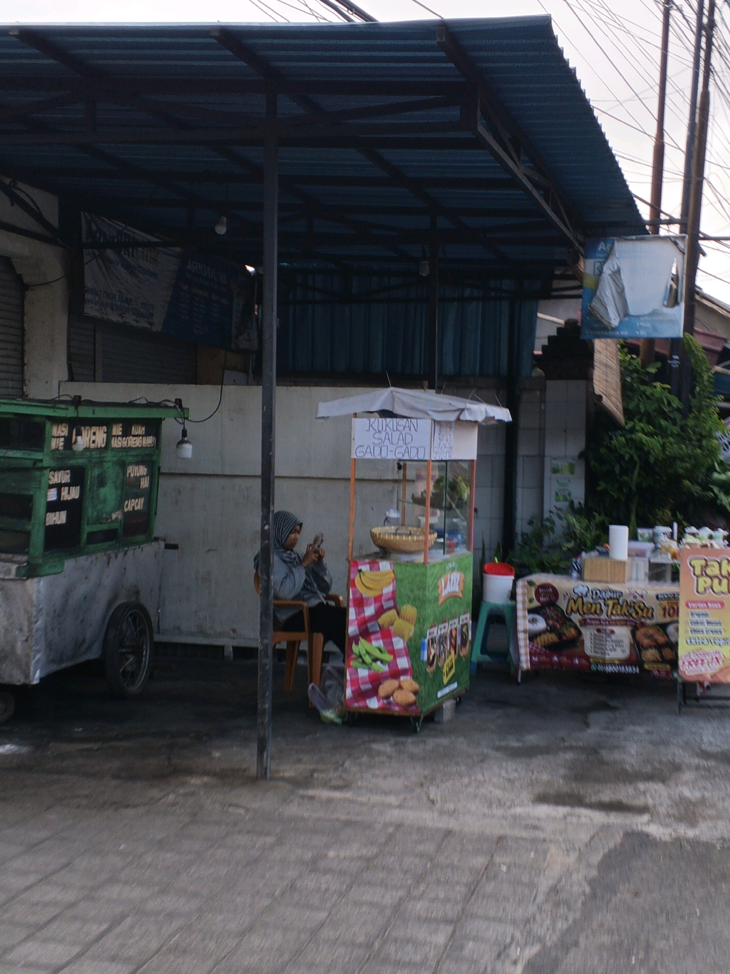 In Denpasar, Indonesia, an outdoor food stall area is depicted. A woman wearing a gray headscarf and jacket sits on a wooden chair to the left, looking at her phone. In front of her is a green food cart with white lettering that lists various dishes such as "NASI MIE," "SAYUR HIJAU," "BIHUN," "GORENG," "KUAH," "NASI GORENG," "PUYUNG HAI," and "CAPCAY." To the right, a smaller food cart with a white canopy displays "KUKUSAN SALAD GADO-GADO." Further to the right, another stall features a sign with Indonesian text and images of food, including "MEN TAK SU" and "TAKE PU..." There are several plastic stools and containers visible. The area is paved with gray cobblestones, and the scene is under a blue corrugated metal awning. Power lines are visible overhead.