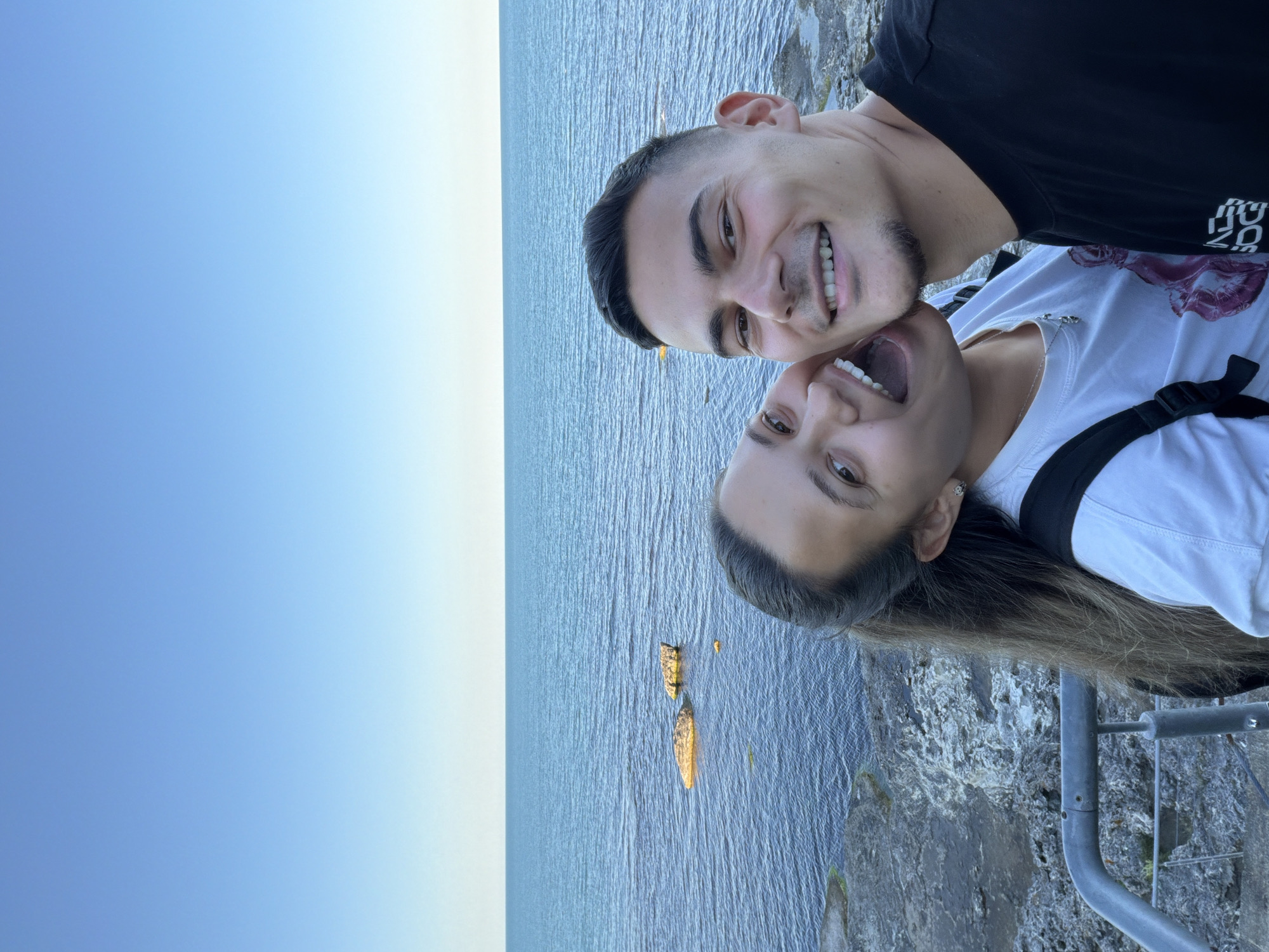 A young couple is taking a selfie on a rocky coastline with the sea in the background. The woman on the left is leaning in towards the man on the right, with her mouth open in a laugh and her head tilted. She has long, brown hair pulled back into a ponytail and is wearing a white t-shirt. The man on the right is smiling and looking at the camera. He has short, dark hair and is wearing a black t-shirt. The sea is a deep blue and there are several rocks visible in the water. The sky is clear and blue. The location is Tsarevo, Bulgaria.