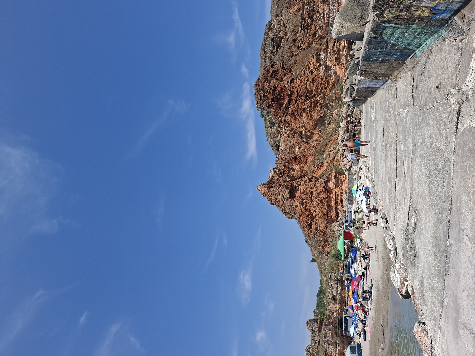 This is a vertical, eye-level, full shot of a sunny beach in Balgarevo, Bulgaria, near the Kaliakra Cape. The scene is dominated by a bright blue sky with thin, wispy clouds. In the mid-ground, a sandy beach is populated with people lounging under colorful umbrellas and several vehicles, including vans and cars. A prominent, rugged, reddish-brown cliff face rises behind the beach. In the foreground, a concrete pier or walkway extends from the right side of the frame towards the left. The pier is marked with graffiti on its side. Several people are walking on the pier, and others are visible on the beach. The water of the sea is visible on the left side of the pier. The overall impression is a lively summer day at a coastal resort.