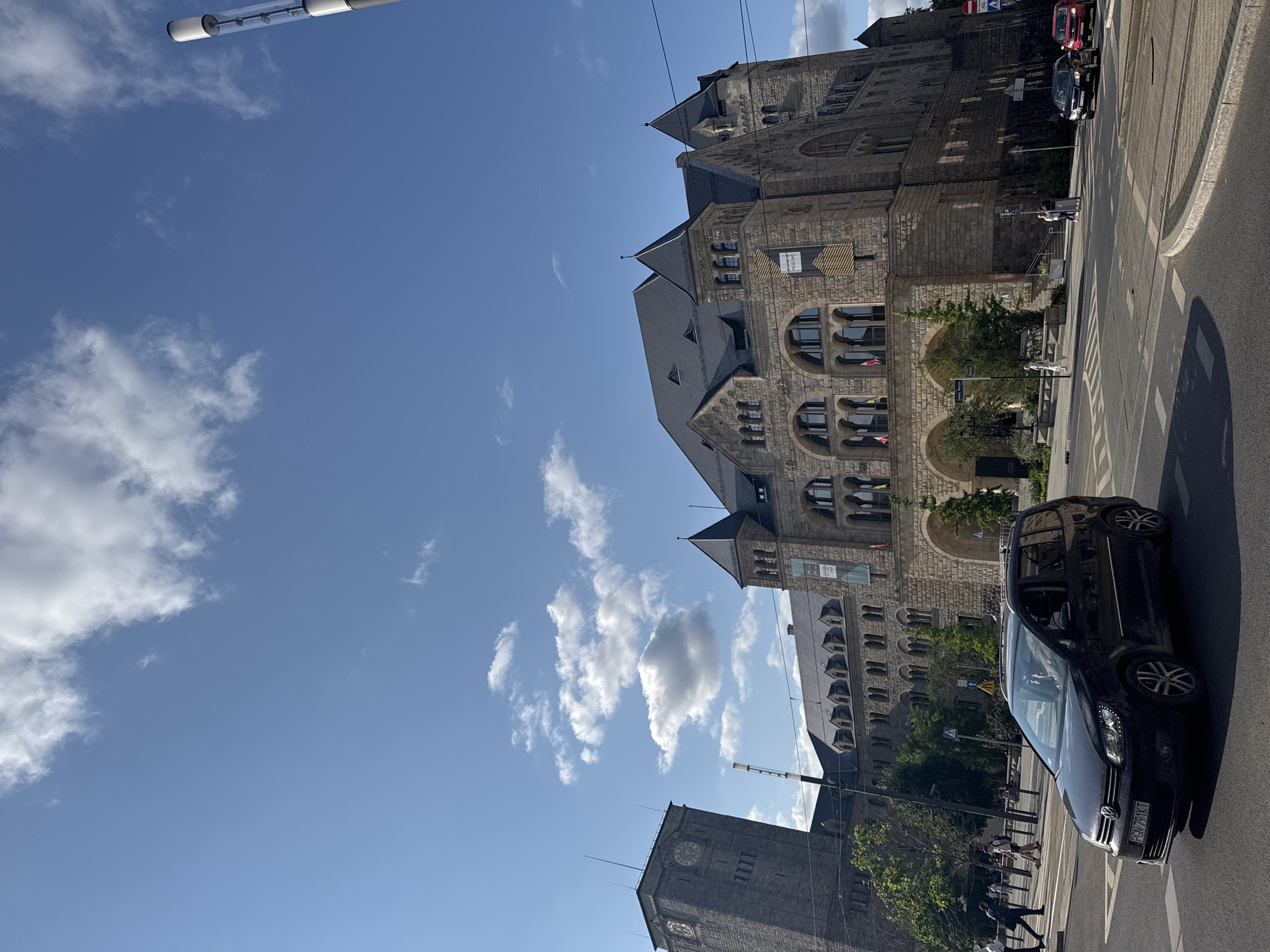 The image shows a view of the Imperial Castle in Poznan, Poland, on a sunny day with scattered clouds. The castle is a large, ornate stone building with multiple towers and dark roofs. In the foreground, a black Volkswagen Touran is driving on a city street, with two red cars visible in the distance. Pedestrians are walking on the sidewalk in front of the castle. A tall, modern street light stands on the right side of the image. The sky is clear blue with white clouds.