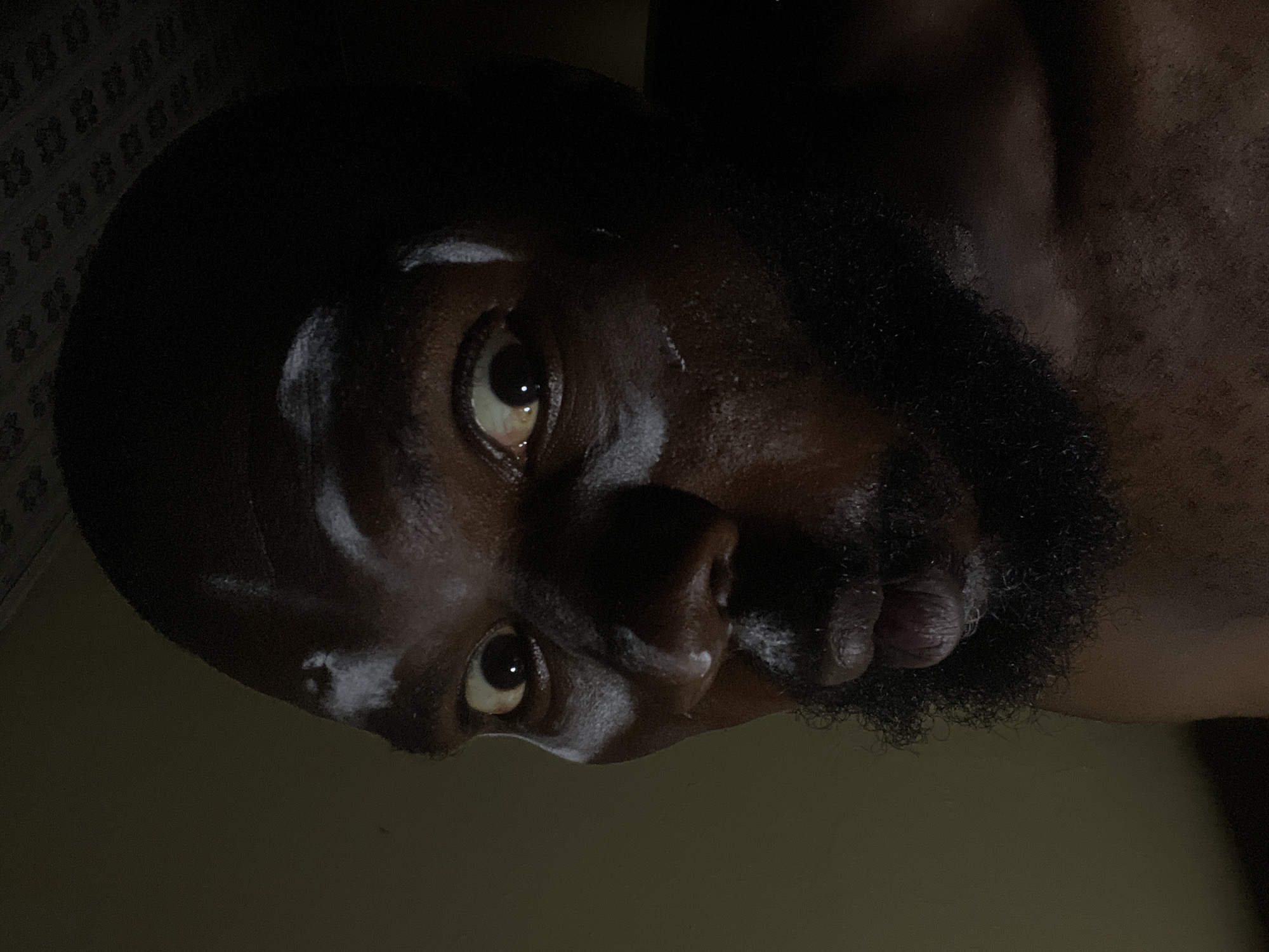The image depicts a close-up, upward-facing view of a Black man's face. The man has dark skin, a beard, and short hair. The background is blurry and dimly lit. It is impossible to verify the location in Gura Top, Nigeria or glean specific actions or interactions from the image.