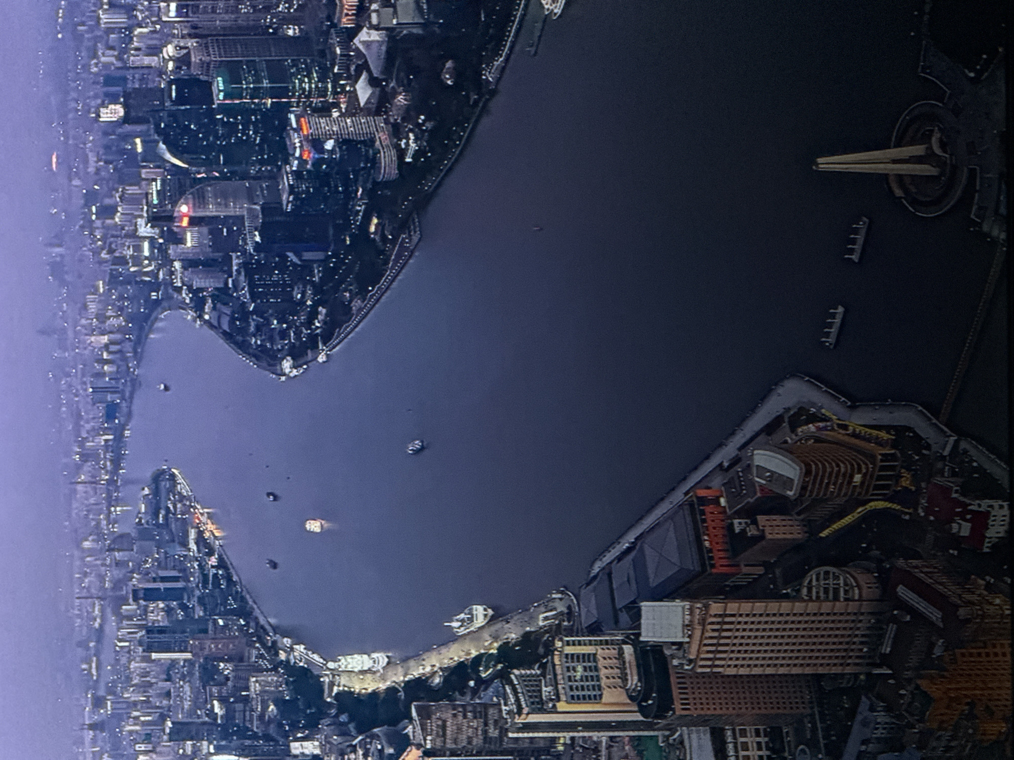 The image shows an aerial view of a city skyline at dusk or dawn, bisected by a wide river. Numerous tall buildings, illuminated by interior lights and some exterior signage, line the riverbanks. Several ships and boats are visible in the water, some docked along the shore and others navigating the river. The overall atmosphere is urban and expansive, with the cityscape stretching into the hazy distance. The scene depicts a bustling port city with modern architecture, as indicated by the density of high-rise buildings. The light suggests it is either early morning or late evening. There is no indication of this scene being located in Saratov, Russia.