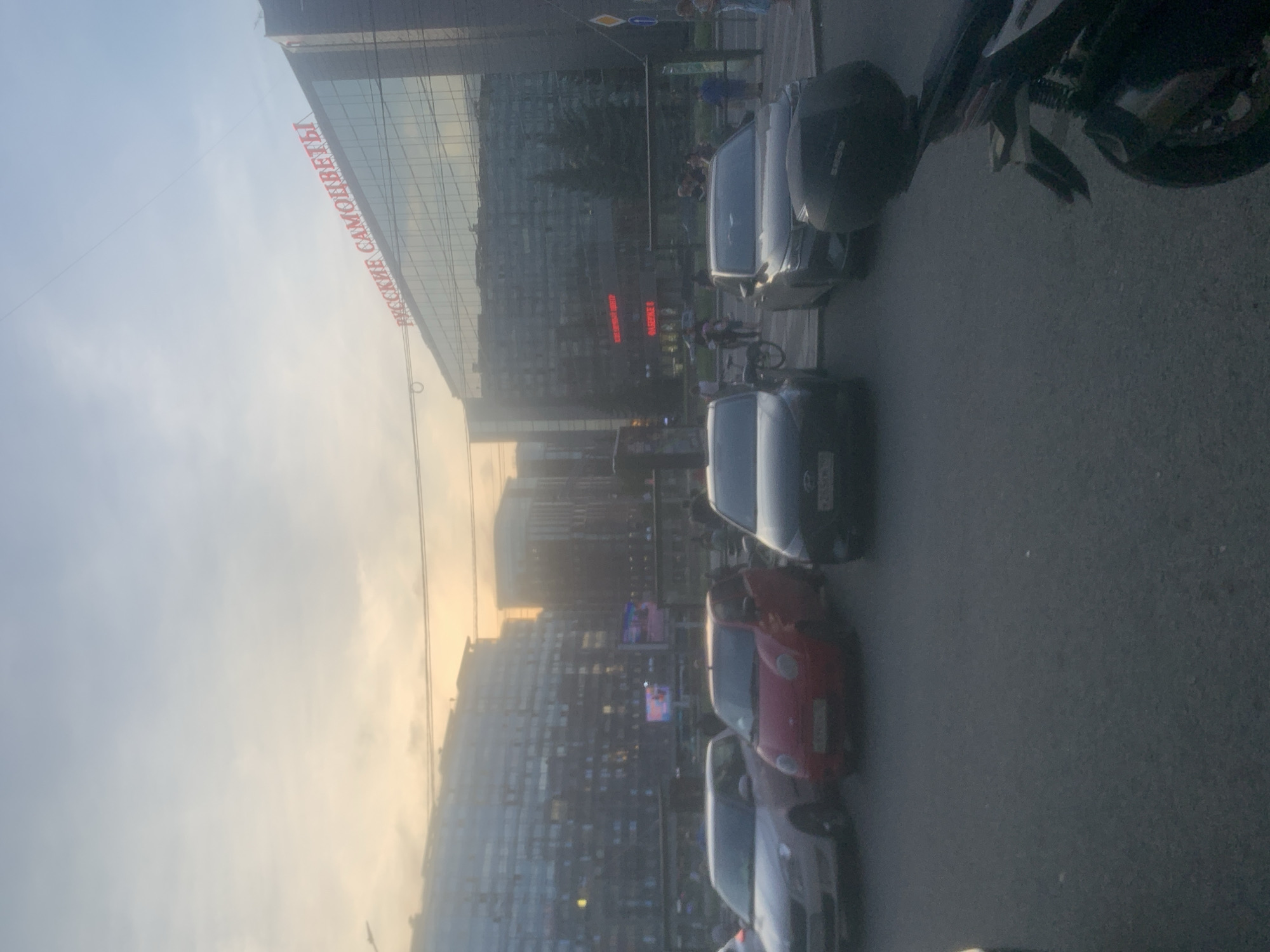 The image depicts an urban street scene in Sinyavino, Russia, bathed in the soft light of either sunrise or sunset. In the background, a large, modern building with a glass facade is prominently featured, bearing the red Cyrillic lettering "РУССКИЕ САМОЦВЕТЫ" (Russian Gemstones). Several cars are parked in the foreground, including a light-colored sedan, a small red hatchback, and two darker sedans. Some pedestrians are visible in the mid-ground, near a bus stop shelter. The environment appears to be a city street with paved roads and sidewalks, and overhead electrical wires are visible against the hazy sky. The overall atmosphere is calm and ordinary, capturing a slice of daily life in the city.