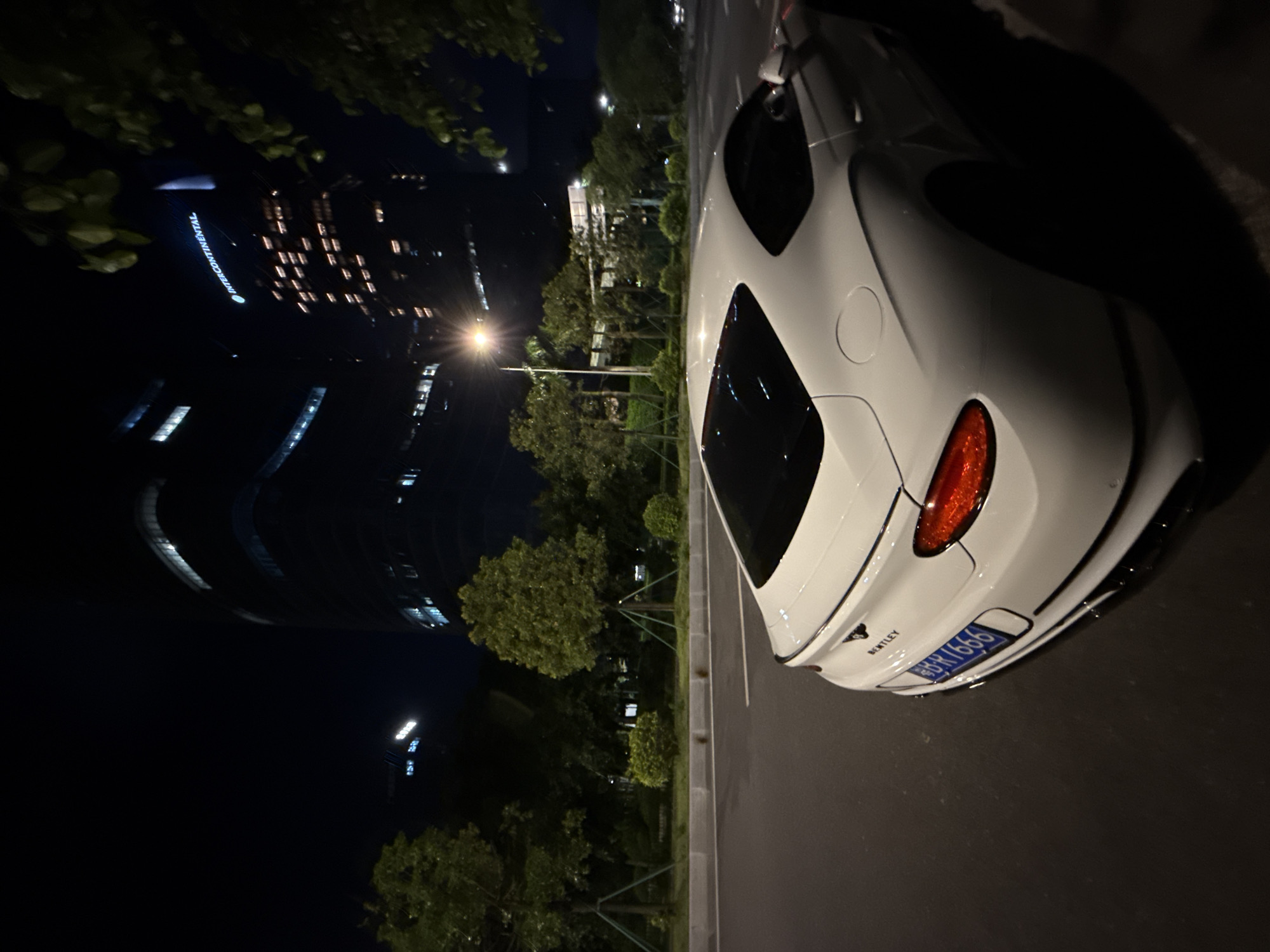 The image shows a white Bentley parked on a street at night. In the background, there are tall buildings, some of which are illuminated. One building has a sign that reads "INTERCONTINENTAL". The area appears to be urban, with trees and streetlights visible. The car is positioned at an angle, with its rear facing the viewer. The license plate is partially visible and appears to be in Chinese. The scene is illuminated by artificial lights from the buildings and streetlights, creating a dramatic nighttime atmosphere. The location is identified as Haizhu District, China.