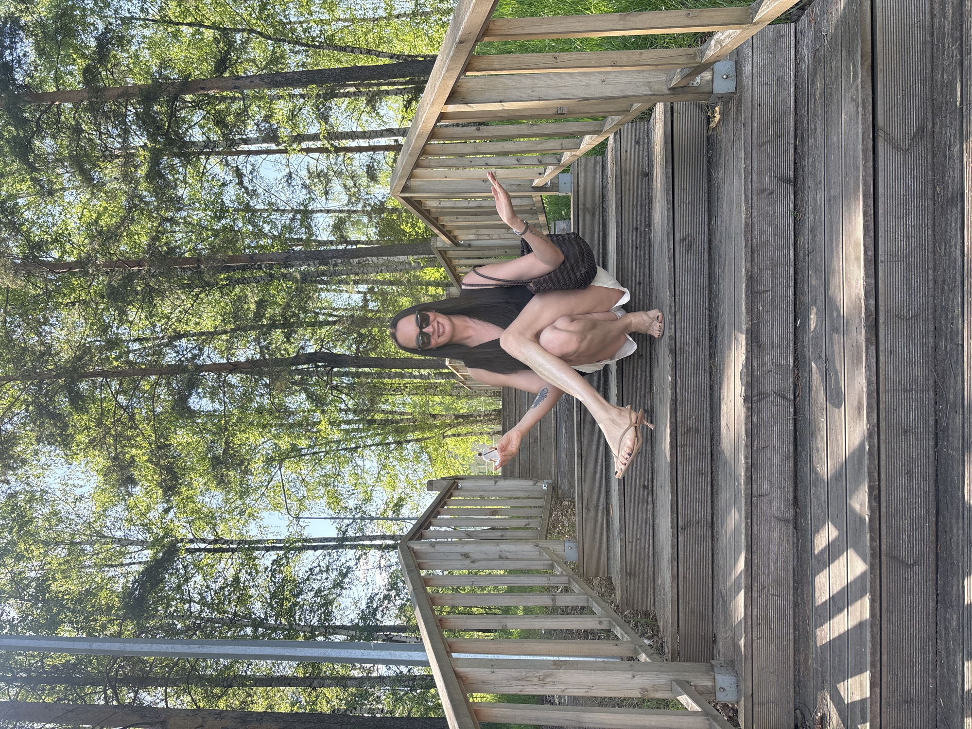 A woman squats on wooden stairs in a park or wooded area in Chaikovsky, Russia. She wears sunglasses, a dark top, light shorts, and heeled sandals, and carries a woven bag. She holds a small white object in her right hand and raises her left hand in a wave. The stairs have wooden railings on both sides, and trees are visible in the background.