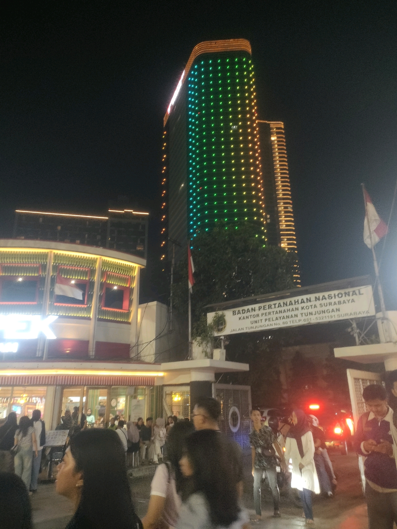 The image depicts a nighttime scene in Surabaya, Indonesia, featuring a modern building facade illuminated by a cascade of colorful lights, predominantly green and yellow, on its upper sections. In the foreground, a group of individuals are visible, some standing and interacting, while others are in motion. One person in the foreground is captured in profile, looking towards the left. Behind the crowd, a sign is mounted on a pole that reads "BADAN PERTANAHAN NASIONAL KANTOR PERTANAHAN KOTA SURABAYA UNIT PELAYANAN TUNJUNGAN JALAN TUNJUNGAN No. 80 TELP. 031-5321191 SURABAYA". A red and white flag is also attached to the pole. To the right of the sign, the taillights of a vehicle glow red. The environment appears to be an urban setting with a focus on a public building and a gathering of people.