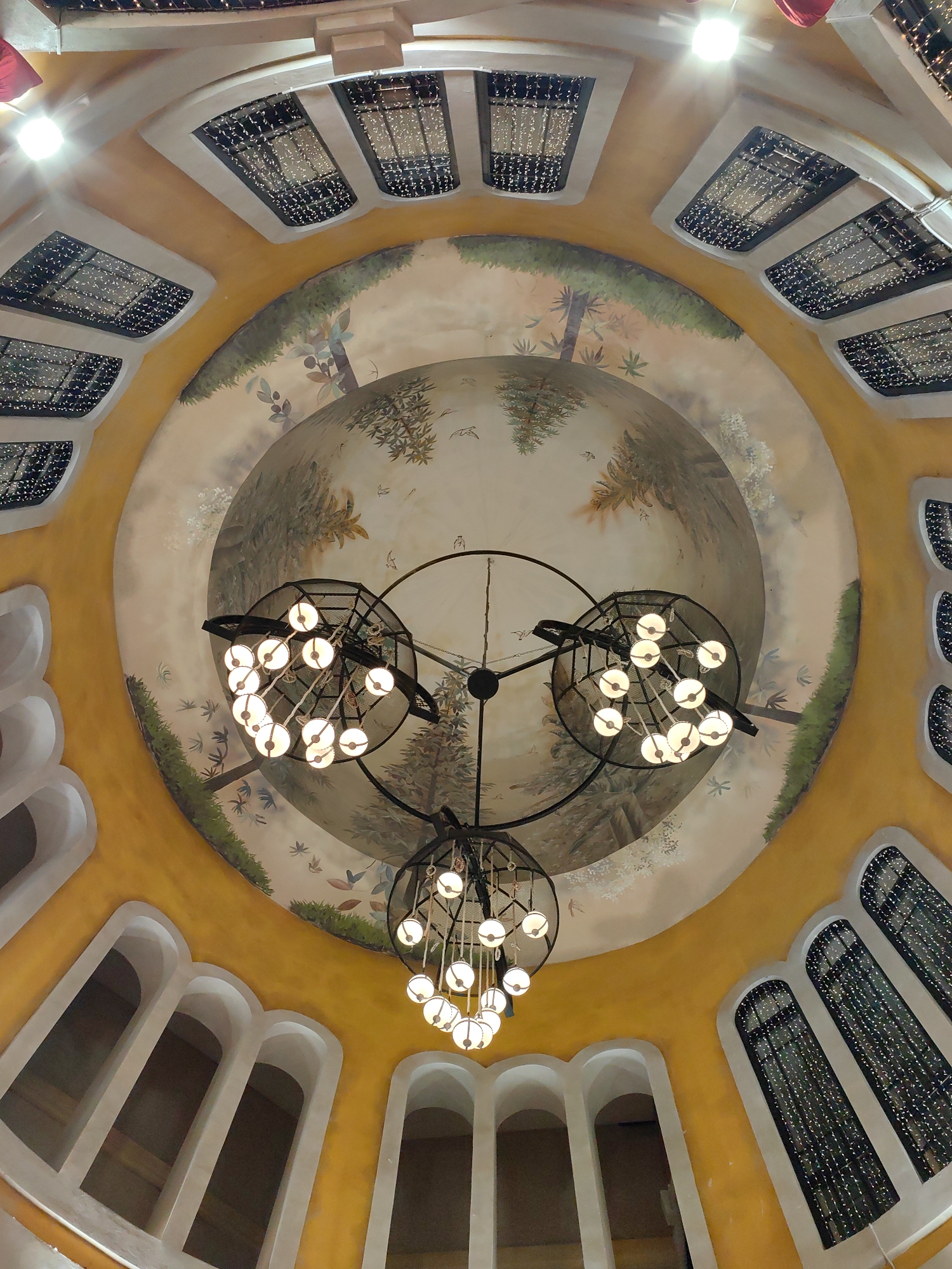 This image presents an upward-looking view of a grand, domed interior ceiling within a building in Sa Pa, Vietnam. The central portion of the dome features a circular mural depicting a natural landscape with various trees, lush foliage, and numerous small, dark bird-like figures in flight against a lighter background. Three multi-bulb chandeliers hang from the center of this mural. Each chandelier has a dark, possibly metallic, spherical or cage-like frame enclosing approximately 10 to 12 illuminated, spherical light bulbs emitting a warm glow.
Surrounding the central mural, the curved architectural walls of the dome are painted a vibrant yellow-orange. These walls are segmented by arched and rectangular openings with prominent light-colored, possibly white, trim. The upper section displays multiple rectangular openings, each filled with what appears to be dense strings of small, white, glittering lights. Below these, larger, empty arched recesses or doorways are visible, maintaining the same light-colored trim. No people are present in the scene.