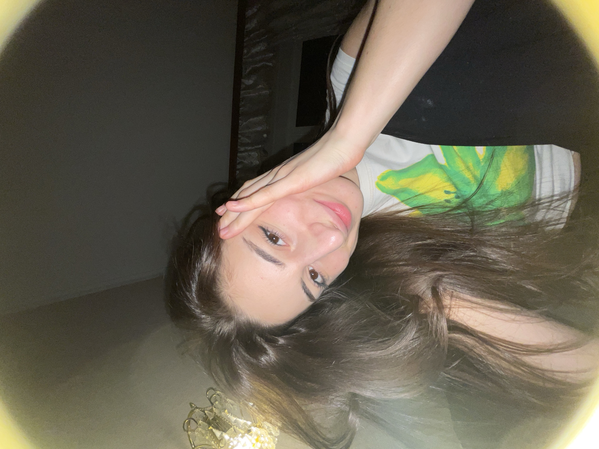 A woman lies with her head near a chandelier. She is wearing a white t-shirt with a large green and yellow design and a black top. Her left hand is on her forehead. The location is unknown and there is no information indicating it is Moscow, Russia.