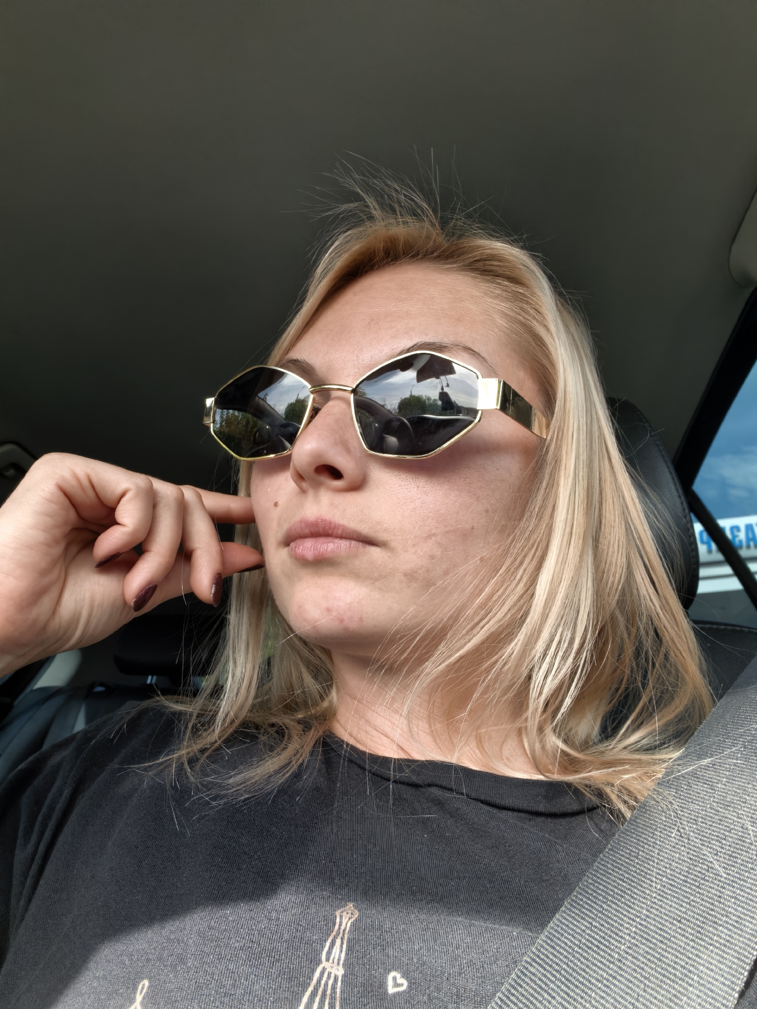 A woman with blonde hair and sunglasses is inside a car, possibly a passenger. She is wearing a black t-shirt and a seatbelt is visible across her chest. Her left hand is resting on her cheek, with her fingers touching her face near her mouth. The interior of the car is dimly lit, with a hint of daylight from the windows. There are no visible signs that this image was taken in Sterlitamak, Russia.