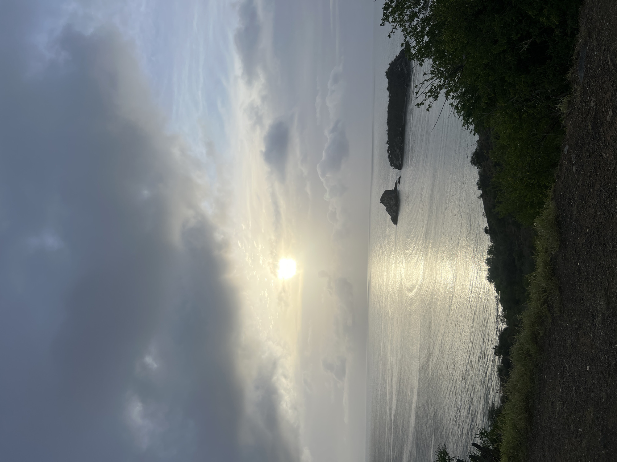 The image shows a tranquil ocean scene under a cloudy sky with the sun peeking through. In the distance, two small islands are visible. The foreground features a dirt path and lush green vegetation. The scene captures a natural landscape, possibly at sunrise or sunset, with the ocean reflecting the sky's light. The overall impression is one of natural beauty and serenity.
The location is identified as being in Manzanillo, Venezuela.