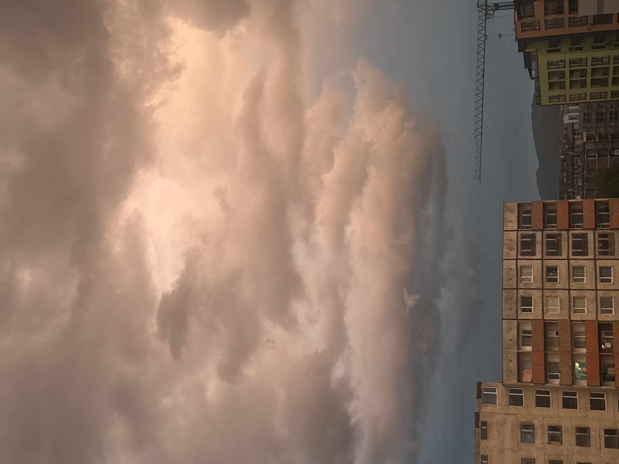 The image depicts a view of Sofia, Bulgaria, featuring a dramatic sky filled with dark, voluminous clouds tinged with hues of orange and gray, possibly at sunset. On the right side of the frame, there are several buildings, with a mix of older, Soviet-era structures exhibiting a grid pattern of windows and newer buildings. A construction crane is visible, suggesting ongoing development in the area. In the background, a faint mountain range can be seen.