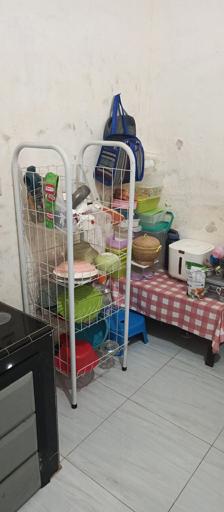 The image depicts a cluttered kitchen or storage area. A white wire shelf unit is filled with various kitchenware, including bowls, containers, and what appears to be a packet of spices. Above the shelf, two bags are hanging on the wall. To the right of the shelf, a table covered with a red and white checkered tablecloth holds a white rice cooker, a water bottle, and other kitchen items. A small blue plastic stool is visible beneath the table. The wall in the background is uneven and shows signs of wear. The floor is tiled. The overall impression is one of a functional but somewhat disorganized living space. The context of Arjasa, Indonesia, suggests this is likely a residential kitchen within that locality.
