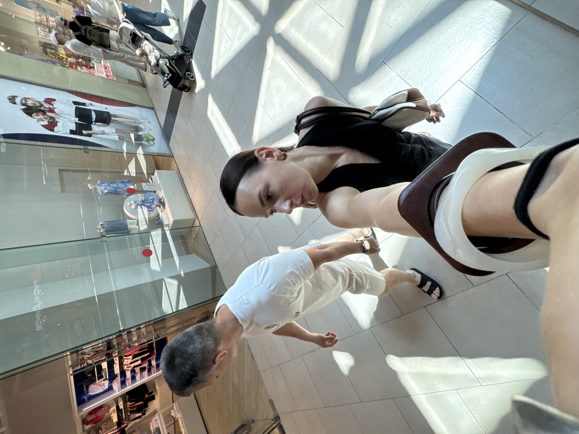 The image presents an interior view of a modern shopping mall located in Sochi, Russia, captured from a slightly elevated, first-person perspective. In the immediate foreground, a female with dark hair pulled back wears a black sleeveless V-neck top and gold-toned hoop earrings, holding the camera with her right arm, while a beige and brown shoulder bag is visible on her left shoulder. To her left and slightly ahead, a male with short gray hair walks, dressed in a white short-sleeved t-shirt, light-colored shorts, and black slide sandals, with a gold-toned watch on his left wrist.
In the mid-ground, further down the tiled corridor on the right, a male figure wearing a white cap and a black backpack pushes a black baby stroller containing a baby, accompanied by a female in light blue jeans and a light top. Other indistinct individuals are visible further in the background.
The mall floor consists of light-colored square tiles, reflecting geometric patterns of strong natural light, indicative of daytime conditions. To the left, a children's clothing store with a glass facade displays various garments in red, pink, white, and blue on shelves, against a prominent blue backdrop. A red circular safety sticker is affixed to the glass, which also shows reversed text reflections. To the right, another store facade features a large promotional poster depicting two young individuals in athletic wear, with a display below showcasing a blue patterned dress on a mannequin and another on a stand.