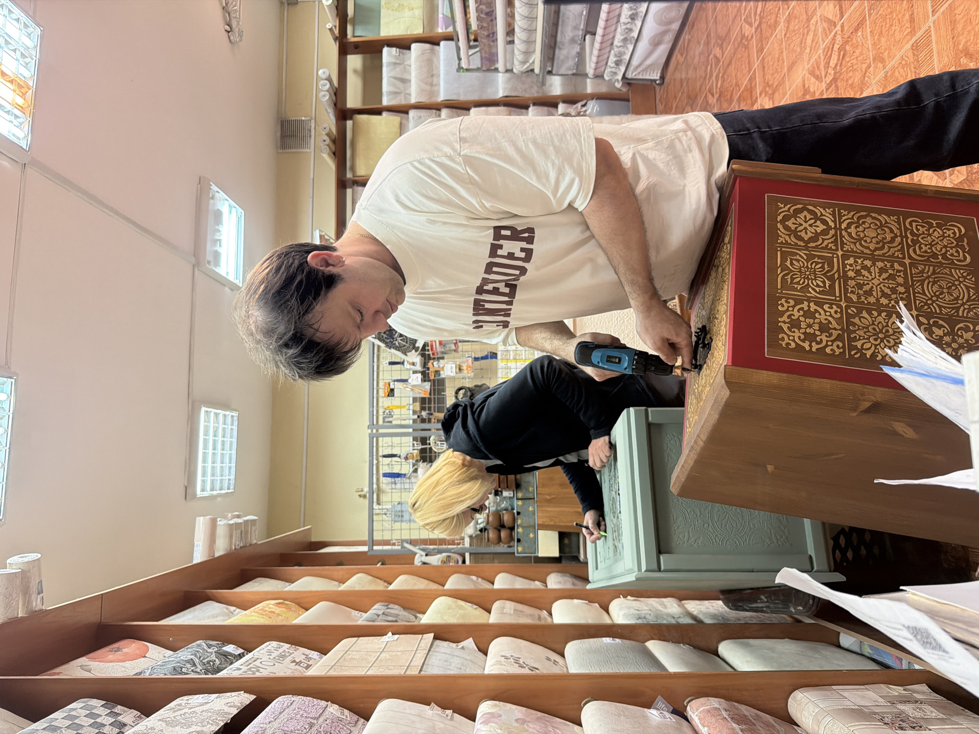 In Maloyaroslavets, Russia, two individuals are engaged in work within a shop, possibly a fabric or wallpaper store, indicated by the numerous rolls of material displayed on wooden shelves lining the left side of the frame. In the foreground, a man wearing a cream-colored t-shirt with maroon lettering that reads "CONTENDER" and dark jeans is using a cordless drill to attach something to a red-and-gold decorative wooden box. His focus is on the task at hand. To his left, a woman with short blonde hair and wearing a black hoodie is bent over a light green chest or cabinet, appearing to be drawing or marking on its surface with a pen. The chest has an embossed or carved decorative pattern. The floor is tiled with orange and brown square tiles. In the background, more shelves with rolls of material are visible, along with some tools and various items stacked on shelves. The lighting suggests an indoor commercial space.