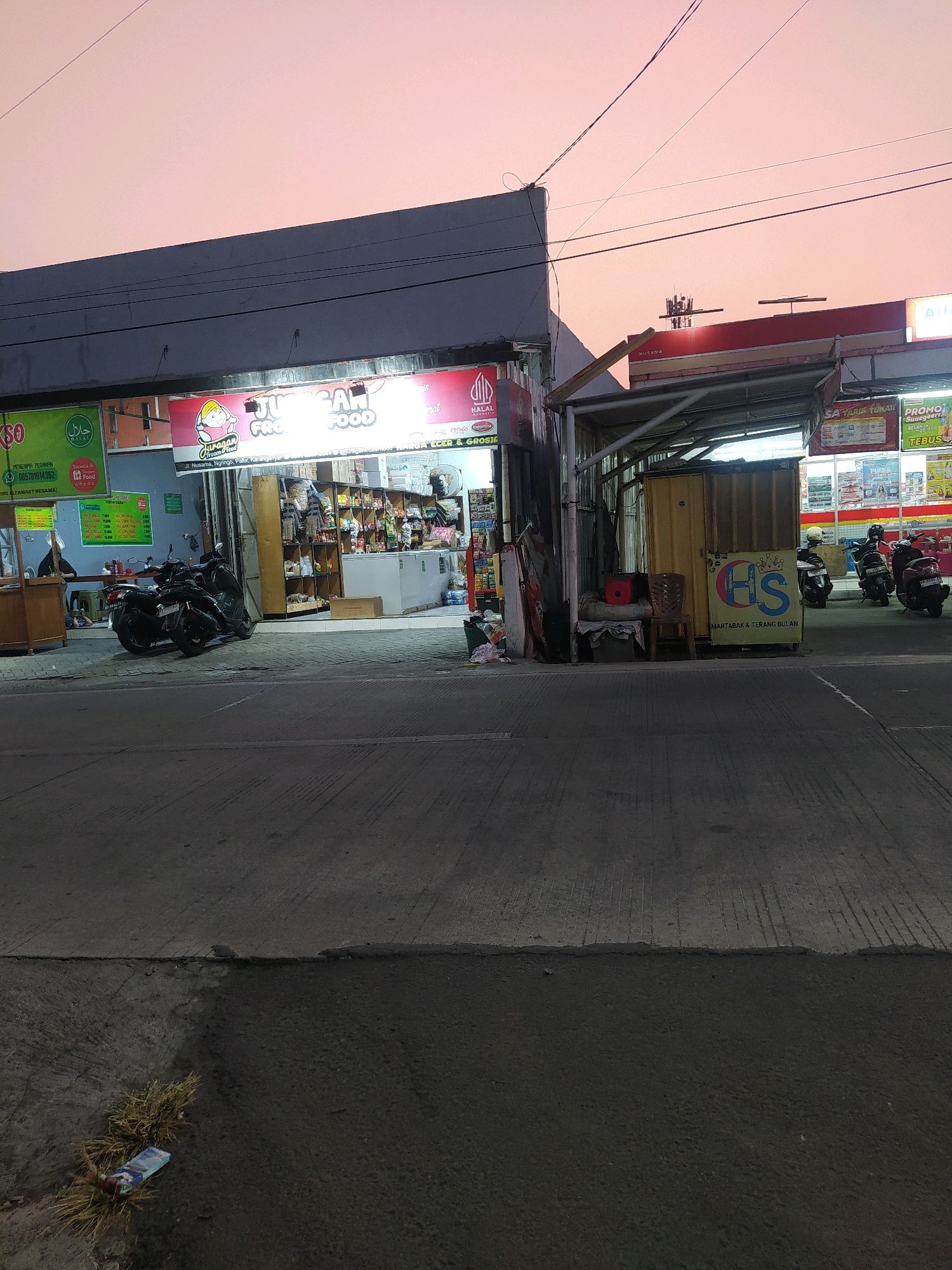 The image depicts a street scene in Jaten, Indonesia, during the twilight hours. The sky is a pale pinkish-orange, indicating either sunrise or sunset. On the left side of the frame, a building houses a shop with a prominent sign that reads "JURAAGAN FOOD". A green sign with a "HALAL" logo is also visible. Two motorcycles are parked in front of this establishment. To the right, another storefront, possibly a convenience store, is partially visible, featuring advertisements and a covered seating area. Several more motorcycles are parked in front of this store, some with riders. A yellow shipping container is placed near the entrance of the second store, with a sign displaying "HS MARTABAK & TERANG BULAN" below a logo. The foreground shows a paved road with a distinct line dividing two different textures of asphalt. Some dry grass and a discarded plastic item are present on the lower left side of the road. The overall atmosphere suggests a quiet evening in a commercial area.