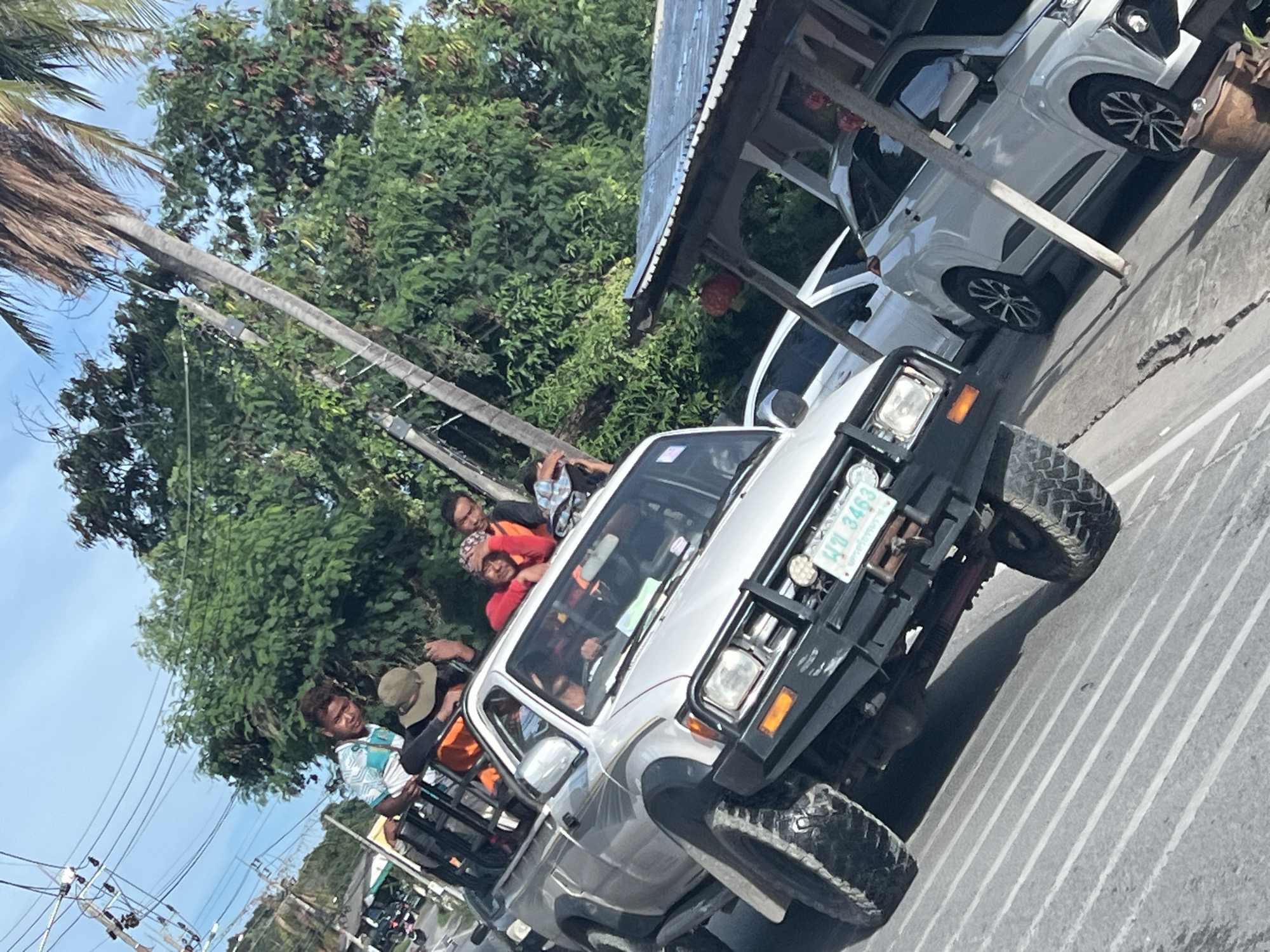 The image depicts a white pickup truck driving on a paved road in Baan Bang Rak, Thailand. Several people are visible in the back of the truck, some in the truck bed and others inside the cabin. A tall palm tree is seen to the left, along with power lines. A building with a metal roof and white vehicles parked beneath it is located on the right side of the road. Lush, green foliage fills the background.