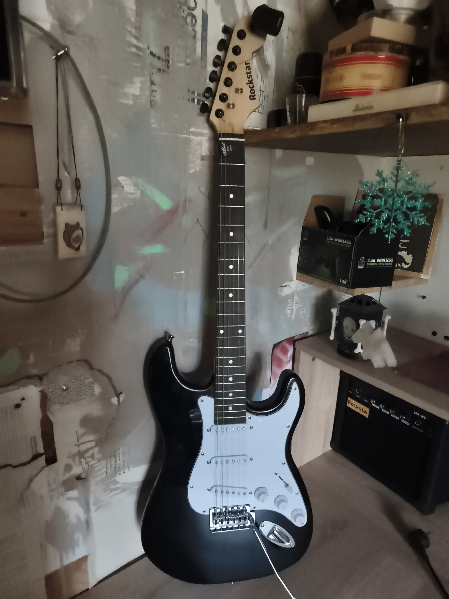 A black electric guitar with a white pickguard stands vertically against a textured, light-colored wall. The guitar has a light wood neck and a black headstock with six tuning pegs. The strings are visible, running down the fretboard which has white dot inlays. The body of the guitar is sleek and curved. To the right of the guitar, on a wooden shelf, sits a small black amplifier with the brand name "Rockstar" visible. Above the amplifier, on another shelf, are various objects including boxes, a green decorative snowflake, and a black lantern-like object with white figures inside. A power cord extends from the amplifier onto the wooden floor. The wall has a distressed, somewhat artistic texture with faint pastel green and pink markings. A hanging decorative item with wooden beads and a cat silhouette is suspended on the left side of the wall. The scene appears to be an indoor setting, possibly a room in a home. The location is specified as Гаранскі сельскі Савет, Belarus.