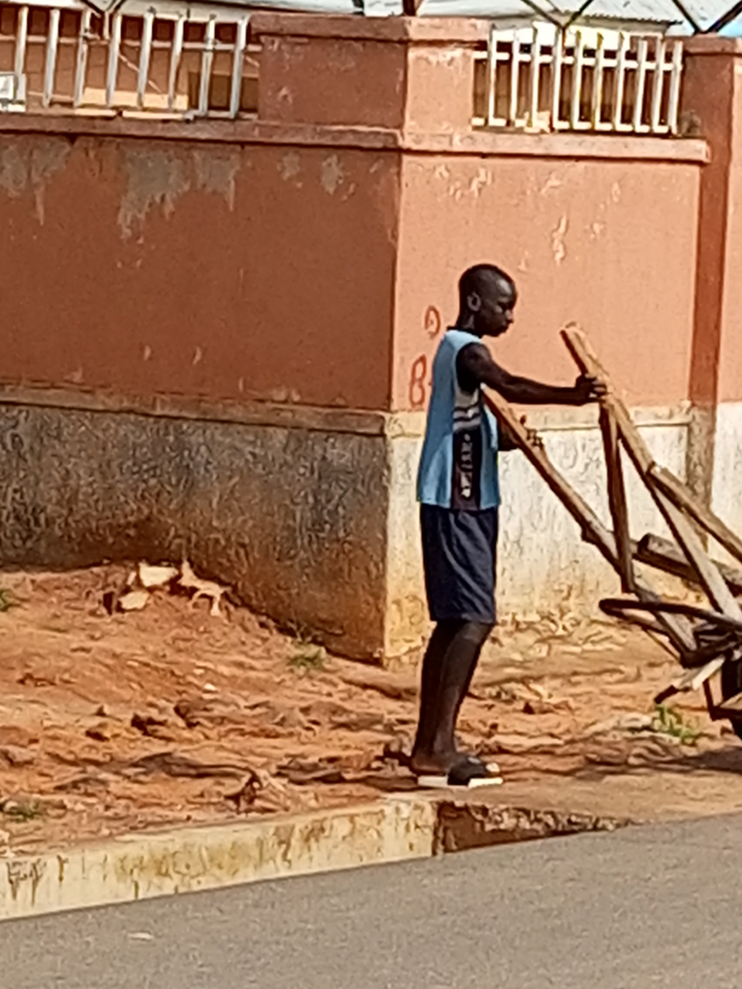 A person stands on a sidewalk in front of an orange-colored building. The building has a white-trimmed window structure at the top. The person is wearing a blue tank top and dark shorts, along with black sandals. They are holding onto a wooden structure that appears to be a cart or a tool with wooden poles. The ground around the person is dirt and rocks. The sidewalk has a concrete edge, and a road is visible in the foreground.