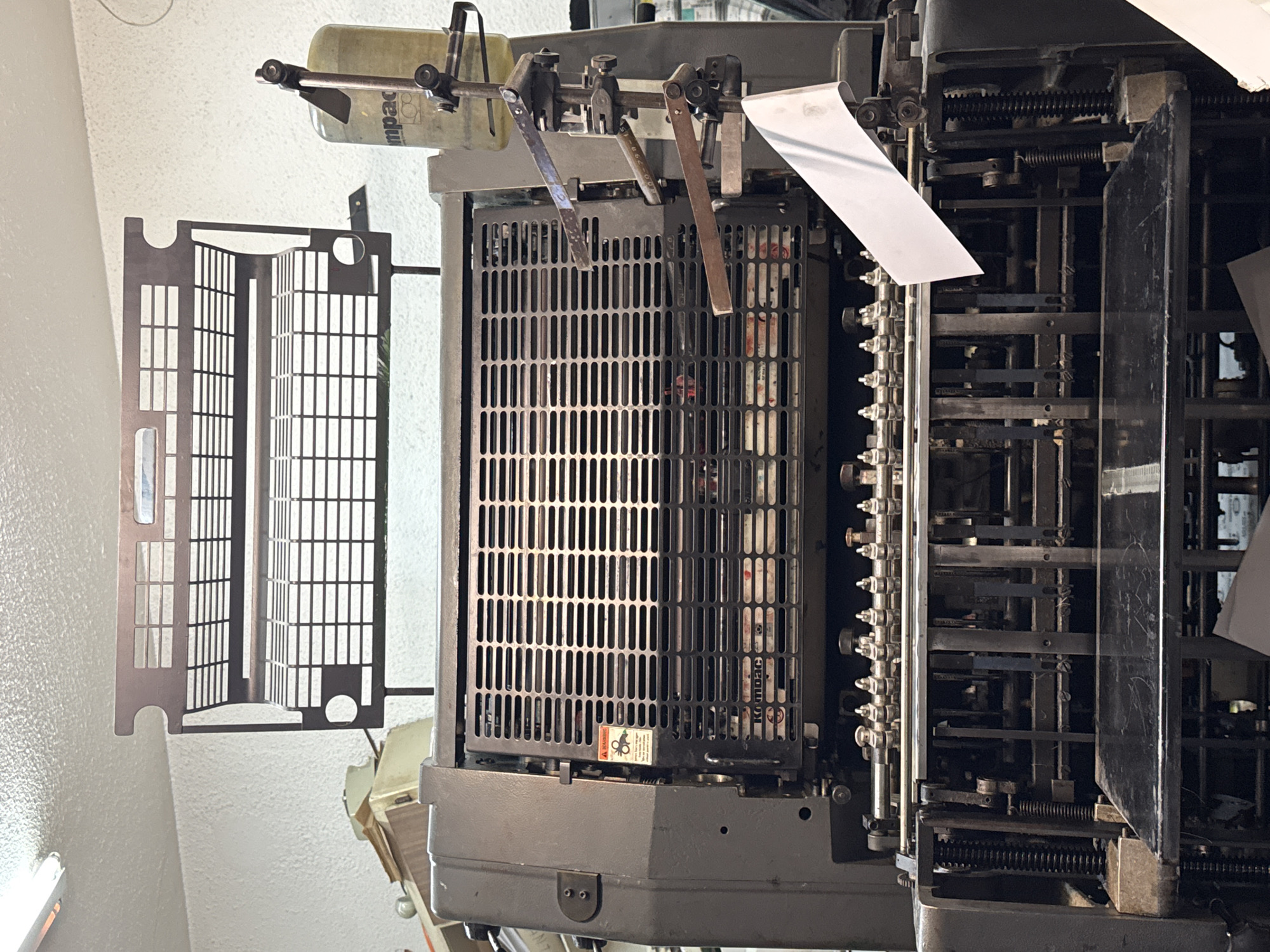 This media file captures a detailed, close-up view of a complex industrial machine, likely a printing press or a similar mechanical device, set against a light-colored wall indoors. The machine is predominantly made of dark-colored metal, showing a robust and intricate design.
In the upper-left area, a translucent container, possibly for ink or fluid, is visible with the partial text "impac" printed on its side. Below this, an array of metallic levers and rods extends horizontally across the machine. A prominent feature is a large, black metallic grille with numerous vertical rectangular openings, serving as a cover for internal components. Faintly visible behind this grille is the word "KODAK". On the lower part of this grille, a small, square warning sticker is affixed, displaying the word "WARNING" and a graphic symbol of a hand caught in gears.
To the left of the main machine body, a separate, lighter-toned metallic panel or cover is visible, featuring a distinct pattern of rectangular cutouts, giving it a grid-like appearance. To the right, a vertical arrangement of numerous small, knurled knobs and connecting rods suggests control or adjustment mechanisms. A piece of white paper or film emerges from the right side of the machine, curling downwards. The machine's internal mechanisms, including springs, gears, and various linkages, are visible through openings and gaps in its structure, indicating its complex functionality.
The machine appears to be stationary, with no activity currently taking place. Its industrial aesthetic, with signs of use and intricate mechanical parts, suggests a functional environment. No people are visible in the image. The lighting appears to be artificial indoor lighting.