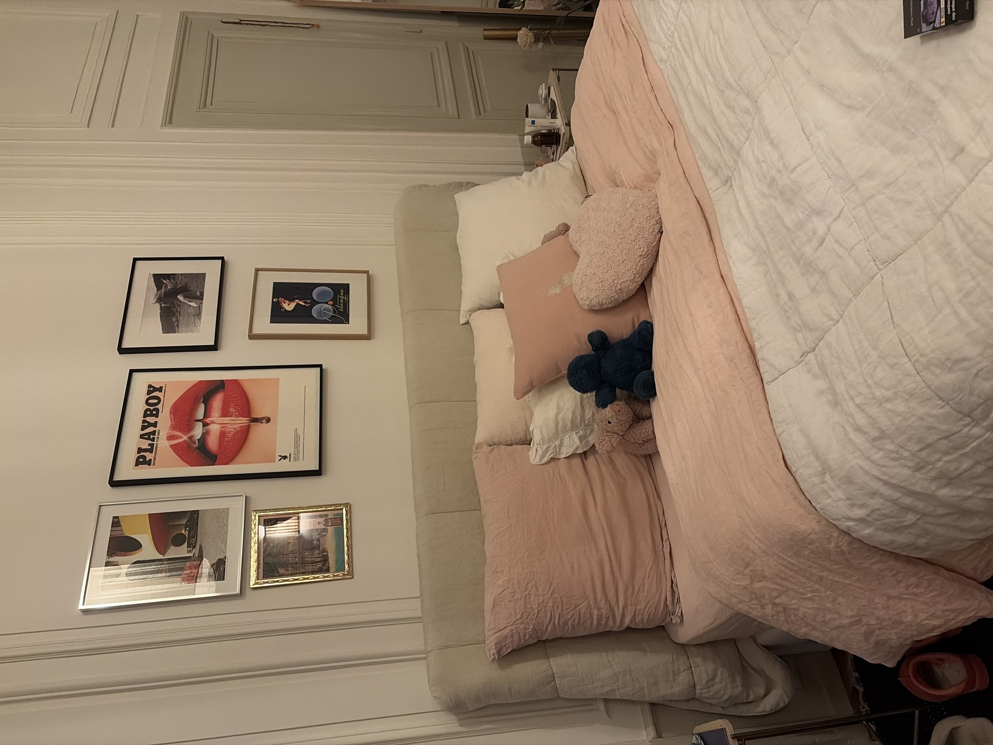 The image depicts an interior view of a bedroom, strongly indicative of a Haussmannian apartment in Paris, France, based on the distinctive classical wall paneling and door design. A bed with a light beige upholstered headboard is centered, dressed with light pink linen sheets and a white quilted comforter. Multiple pillows are arranged, including large crumpled pink ones, white pillows, and a smaller pink one with a visible "Polo Ralph Lauren" tag. On the bed, three plush items are present: a dark blue stuffed toy, a light pink stuffed pig, and a pink heart-shaped pillow.
Above the headboard, five framed pictures are mounted on the white wall. From left to right: a silver-framed photograph of modernist architecture; a smaller gold-framed picture beneath it; a prominent black-framed "PLAYBOY" poster featuring red lips with a burning matchstick; a black-framed black and white photograph of a woman on a beach; and a gold-framed "Pétanque" poster.
To the right of the bed, a light-colored bedside table holds a bottle of La Roche-Posay product and a white mug. A light grey paneled door is visible further right, alongside a partial view of a tall mirror and a decorative object, possibly a vase. On the floor near the left side of the bed, a red slipper and a white slipper are present on a dark floor covering. A pack of Marlboro Red cigarettes rests on the white comforter in the lower right foreground. No individuals are depicted in the scene.