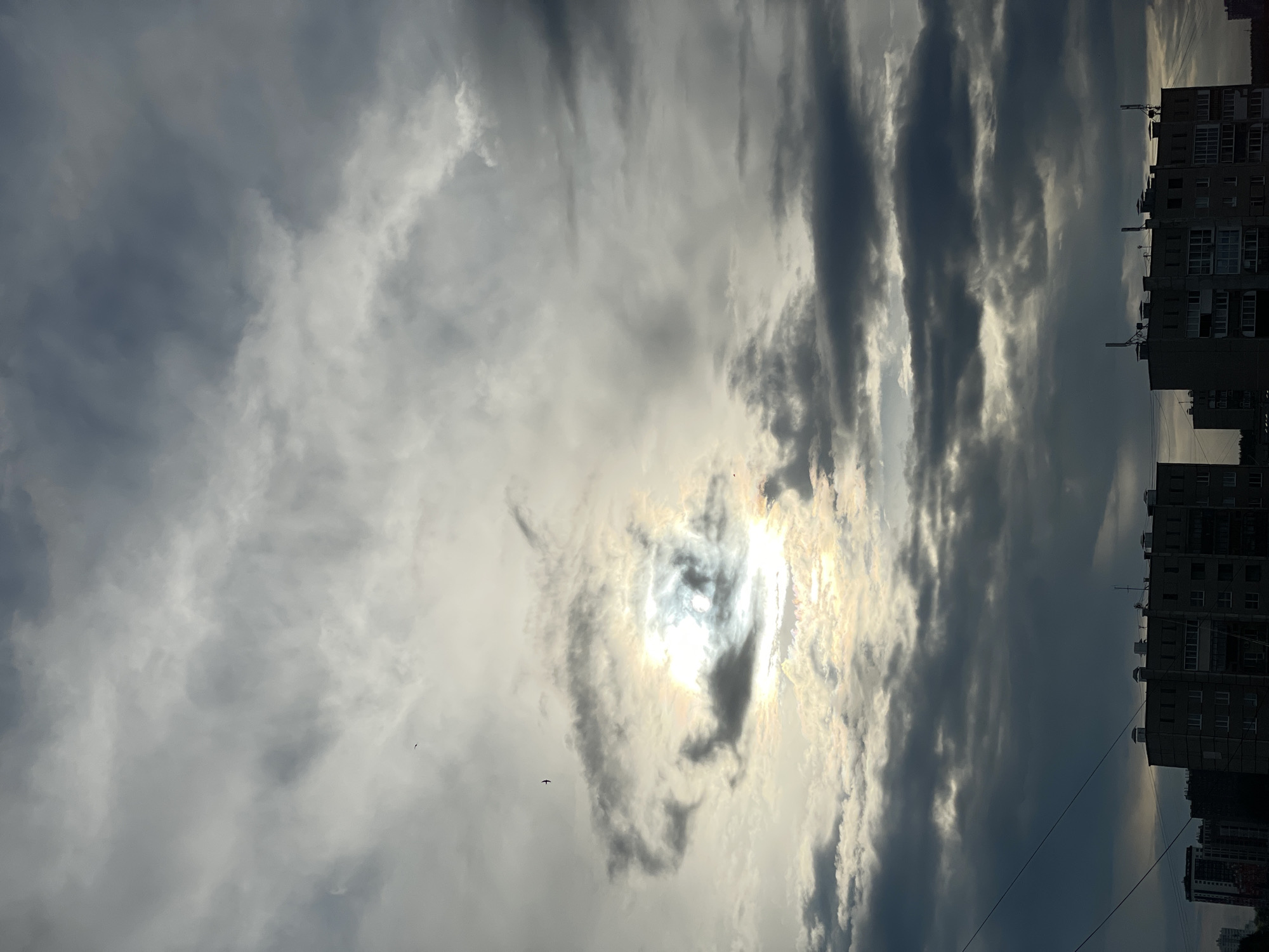 The image displays a dramatic skyscape over an urban setting in Izhevsk, Russia, likely during late afternoon or early evening. The sky is predominantly filled with dynamic cloud formations, featuring a striking contrast between bright, illuminated areas and dark, heavy clouds. The sun, positioned in the upper-middle, shines intensely from behind a veil of darker clouds, creating a powerful halo effect and visible sunrays that pierce through the cloud cover.
Along the horizon, which occupies the lower portion of the frame, a series of multi-story apartment buildings are silhouetted against the bright sky. Their architectural style suggests a post-Soviet era design, common in Russian cities. These buildings appear dense and residential, with visible windows and rooftop structures. Several small birds are captured mid-flight against the lighter, cloudy backdrop, adding a sense of scale and life to the vast sky. No people or visible text are present in the scene. The overall impression is one of powerful natural beauty juxtaposed with an urban landscape, characterized by strong light, shadow, and a dramatic cloudy atmosphere.