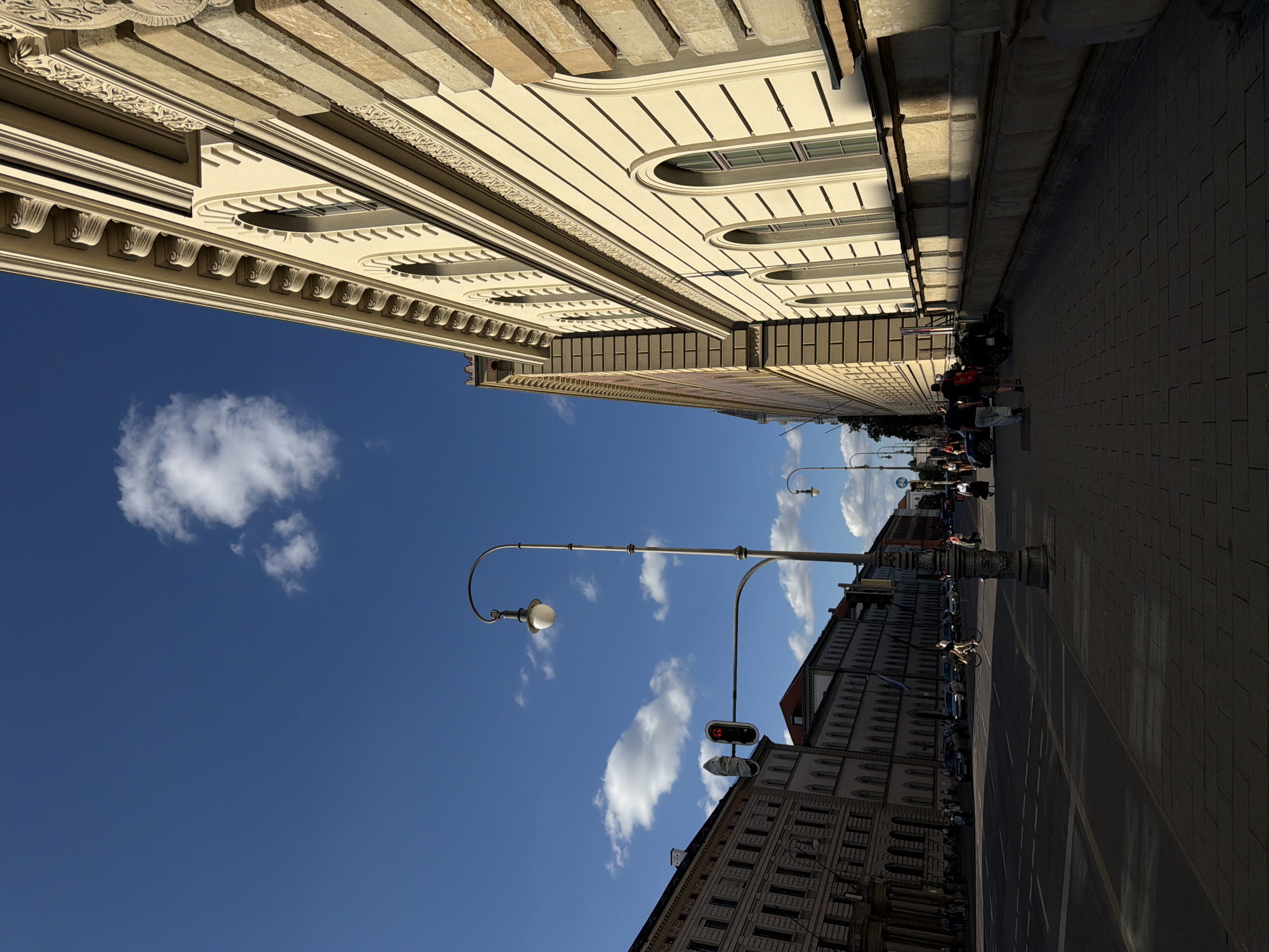 This is an outdoor, eye-level shot of a street in Munich, Germany, with a strong perspective from below, looking up at grand buildings and a bright blue sky dotted with white clouds. The image is oriented vertically, creating a dramatic upward sweep.
The primary subjects are the ornate architectural facades of large, old buildings lining a pedestrian walkway and street. The building on the left is light-colored, with detailed cornices, decorative carvings, and arched windows. The building on the right is also grand, with many windows and a darker roofline visible further down the street.
On the pedestrian walkway, there are several people walking, some with bicycles. A motorcycle is parked near the building on the left. Cars are visible on the street, and a cyclist is riding down the street, casting a long shadow. A street lamp with a distinctive curved arm and globe-shaped light fixture stands prominently in the sky, along with a traffic light showing a red arrow.
The scene appears to be set on a sunny day, likely late afternoon or early evening, given the long shadows cast by the buildings and people. The weather is clear and pleasant. The architecture and general ambiance suggest a European city setting, consistent with Munich. There is no visible text in the image. The overall mood is one of urban activity on a fine day.