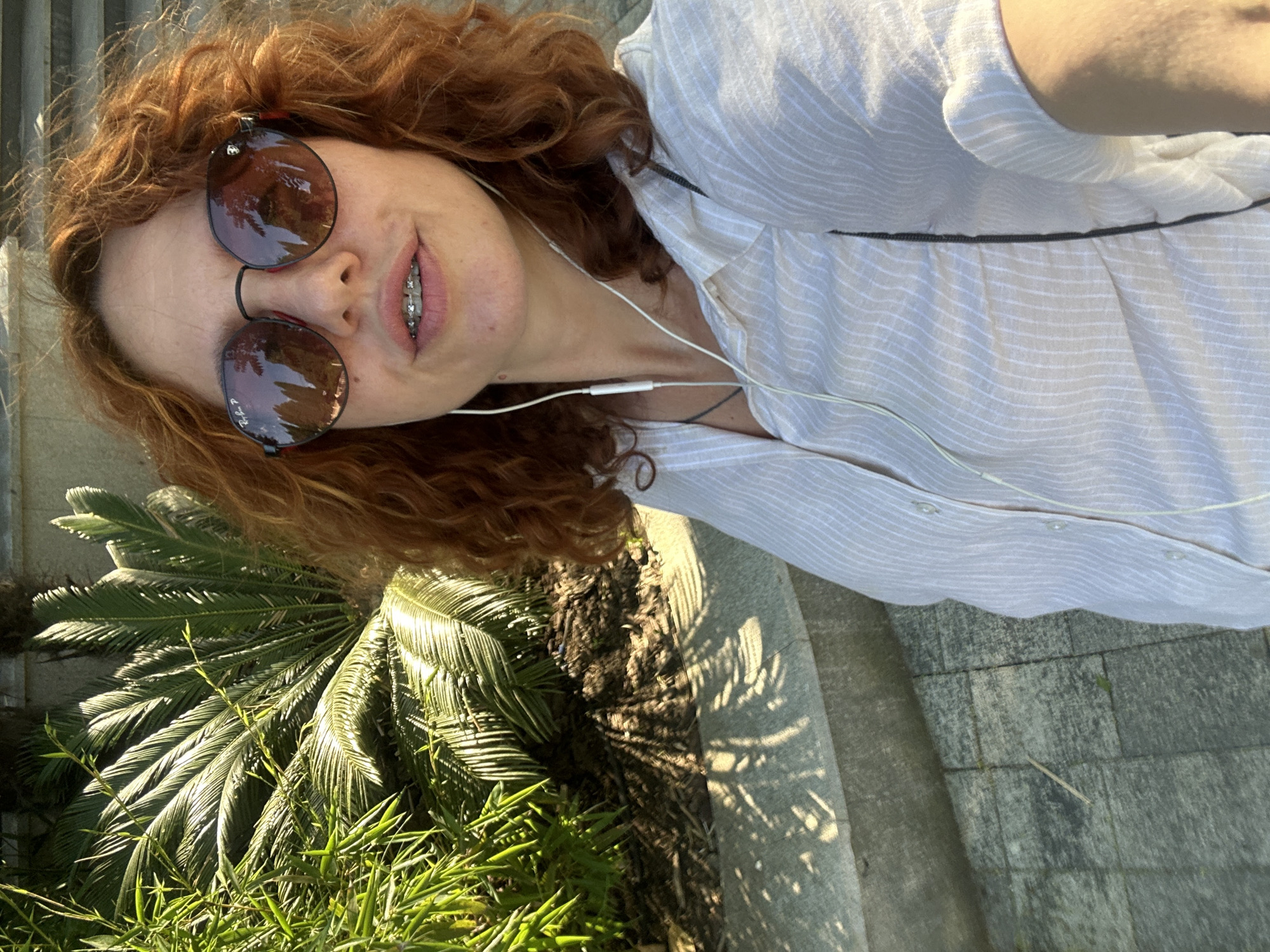 A woman with curly red hair and braces is taking a selfie outdoors. She is wearing round sunglasses and a white striped button-down shirt. She has earbuds in, and the white cord is visible. She is standing in front of lush green foliage, possibly cycad and bamboo plants, with sunlight casting dappled shadows on a stone pathway and ledge. The background suggests an urban park or garden setting in Sochi, Russia, characterized by tropical-looking plants and paved walkways.