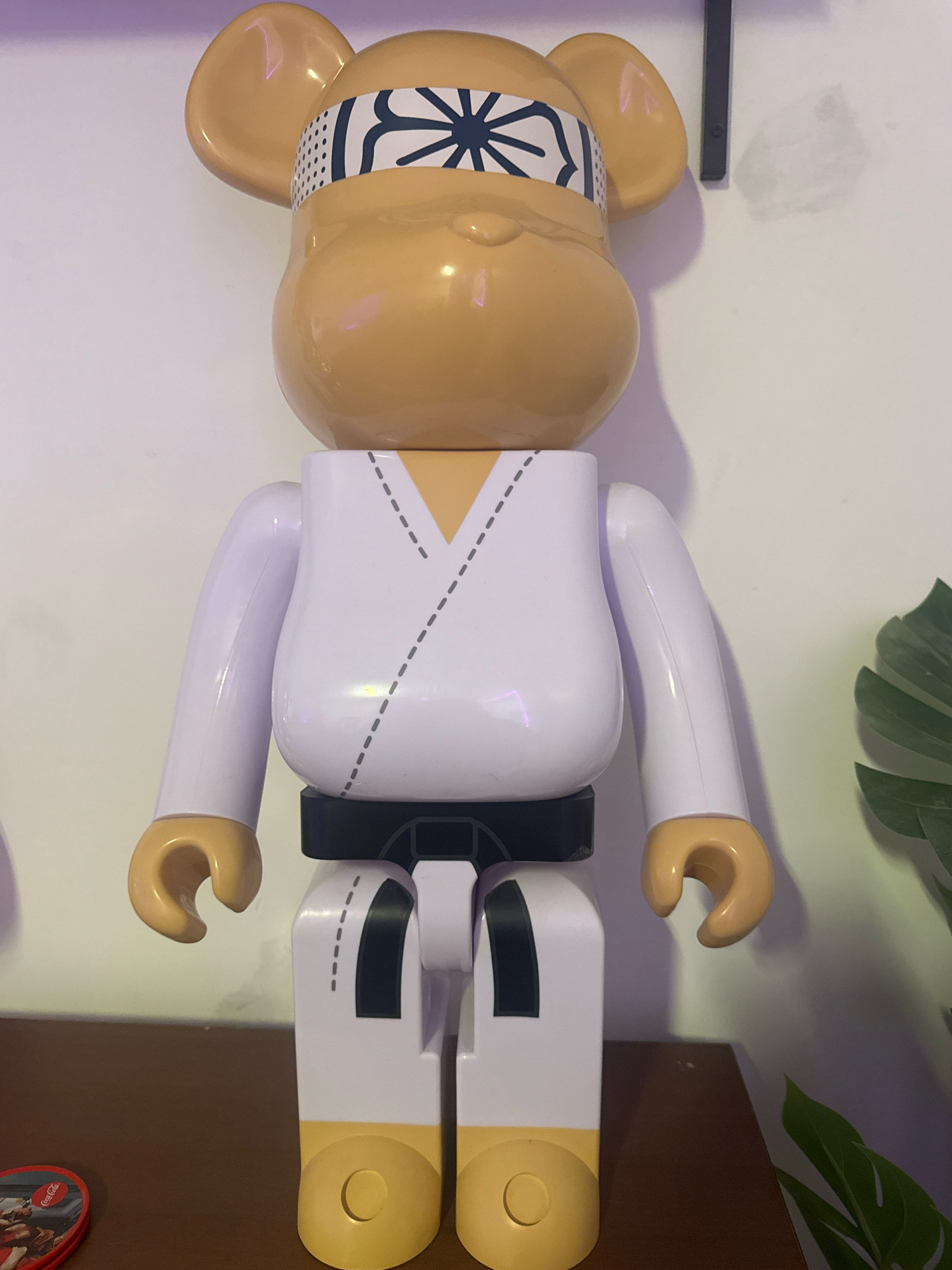 A tan, cartoonish bear figurine, dressed in a white karate gi with a black belt, stands on a brown surface. The figurine has a blue and white headband with a starburst pattern. The background is a white wall with a dark vertical object on the right side. A green leaf is visible to the right of the figurine. The scene appears to be indoors, likely in a residential setting within Germasogeia Municipality, Cyprus.
