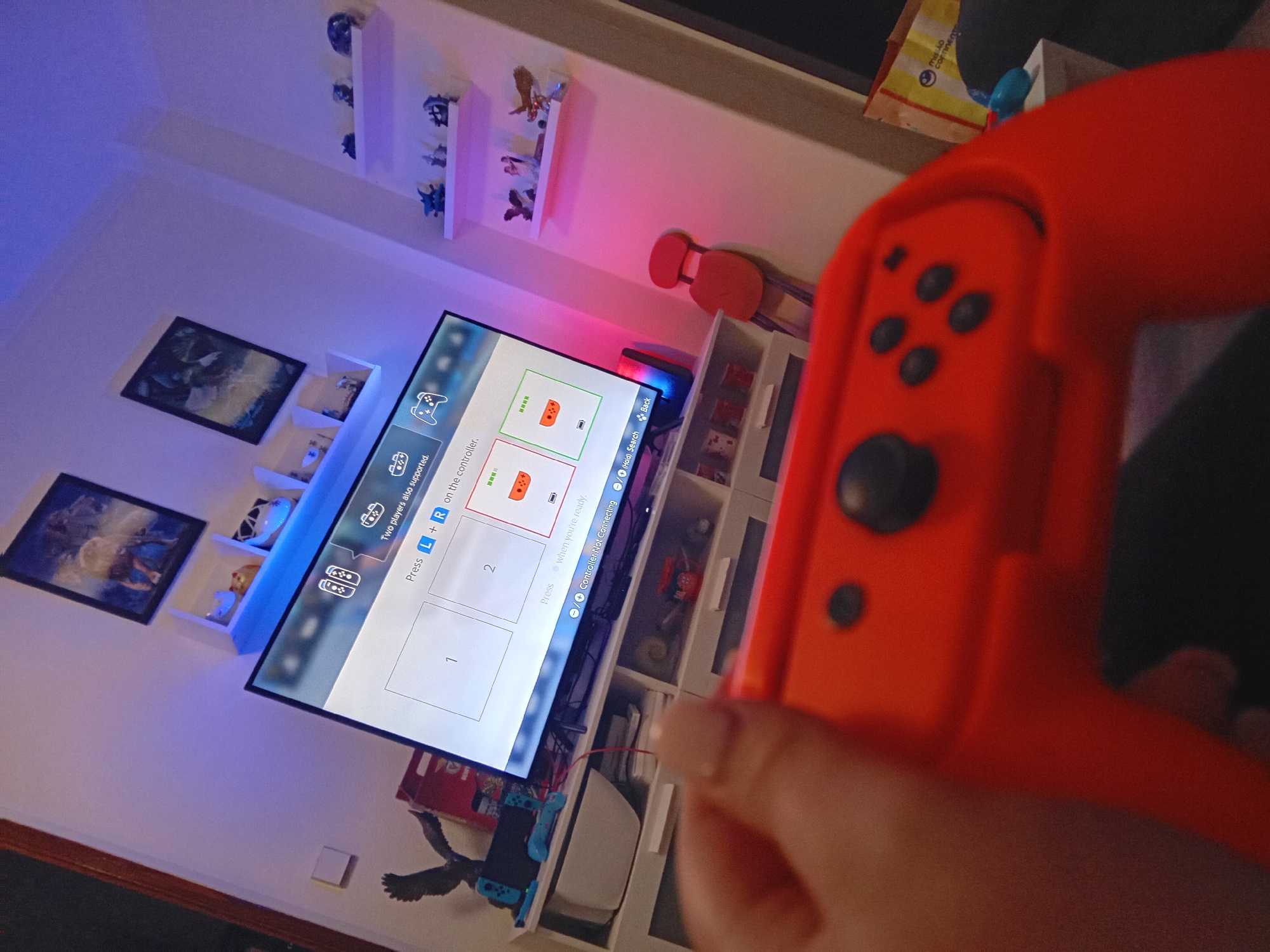 A slightly angled indoor shot features a person holding an orange Nintendo Switch Joy-Con controller in the foreground, while in the background there is a TV displaying a menu for controller pairing. The TV is positioned against a white wall with decorative shelves adorned with small figurines, artwork, and framed pictures. One of the shelves features a small red chair. A white storage cabinet with open compartments is beneath the TV, and some compartments contain various items, including packaging with "MISSÃO CONTINENTE" text. The lighting in the room appears to be provided by LED strips, casting blue and pink hues on the wall. The focus is on the interaction with the gaming console and controller.