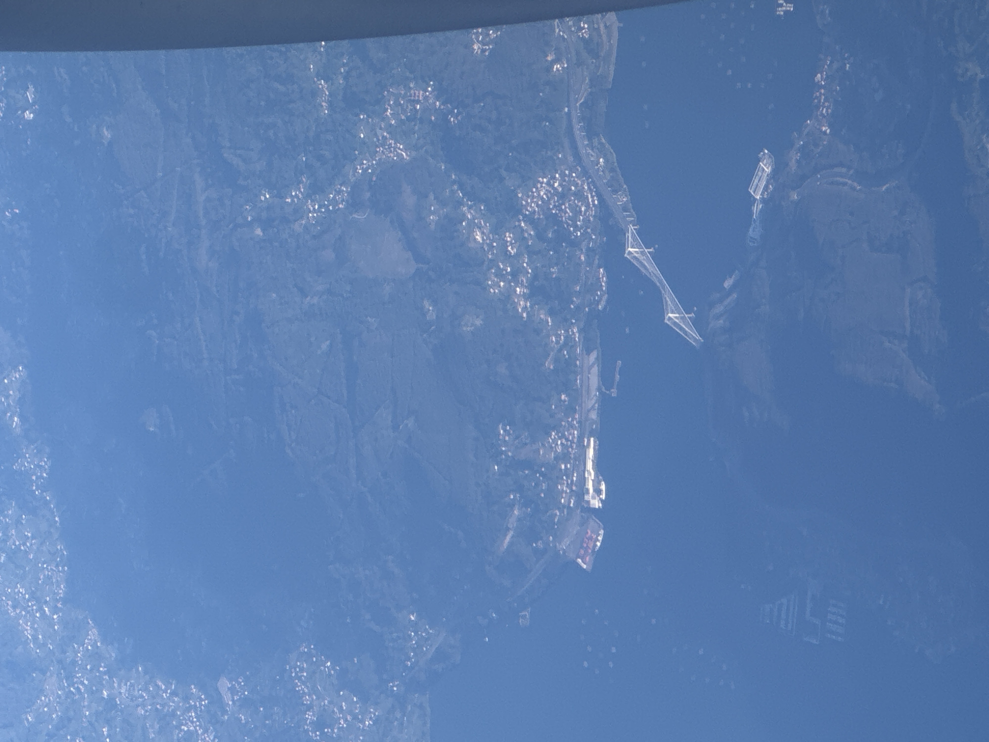 The image shows a coastal area with land on the left and water on the right. The land contains vegetation and scattered buildings that look like houses. A long bridge extends from the land into the water. On the right of the bridge, there is a cluster of rectangular structures or buildings in the water. Further away, towards the bottom-right, there's a larger group of similar buildings.
Without more information, it's challenging to pinpoint the exact location within Sosnowiec, Poland. However, the presence of a bridge, water, and built-up areas suggests a location on the edge of the city near a river or a lake. Given that Sosnowiec is not directly on the coast, this image may not be of Sosnowiec.