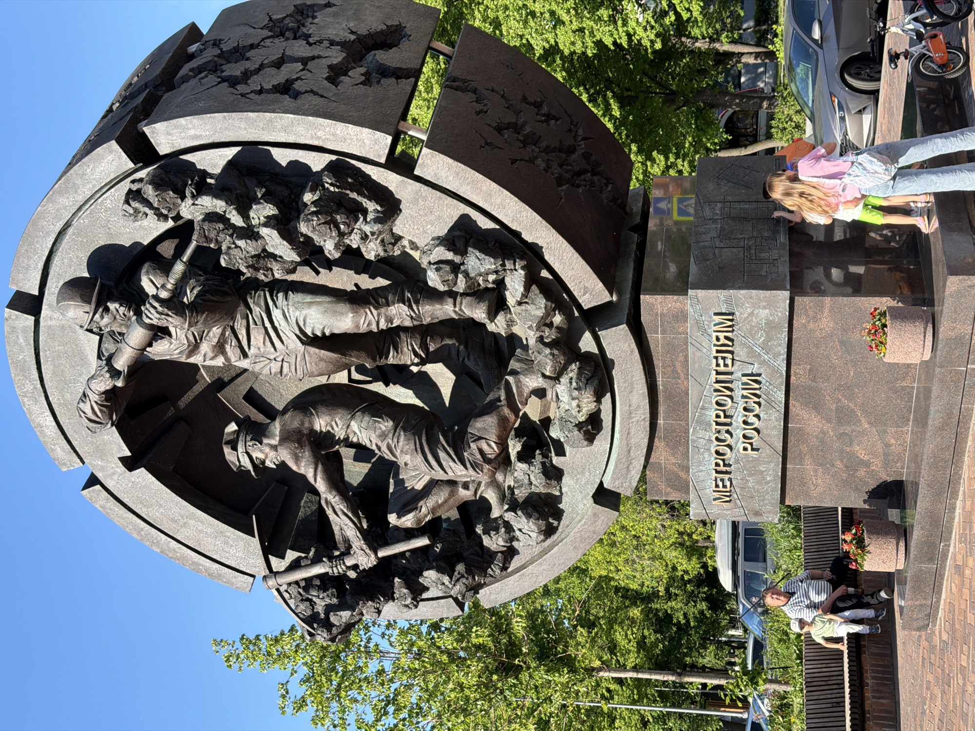 The image displays a bronze monument in Moscow, Russia, dedicated to metro builders, under a clear blue sky. The central sculpture features several dynamic figures of laborers, depicted in action. One figure operates a jackhammer, another wields a pickaxe, appearing to excavate rock and construct a tunnel. The monument is shaped like a partial sphere or tunnel cross-section, with its upper segments intricately designed to resemble fractured earth, symbolizing underground construction.
The sculpture rests on a multi-tiered base made of dark, polished stone. A prominent inscription in Cyrillic reads: "МЕТРОСТРОИТЕЛЯМ РОССИИ," which translates to "To the Metro Builders of Russia." Another section of the base shows an etched diagram, resembling a subway map or architectural blueprint. An adult woman and a child are seen closely examining this diagram. In the foreground, a man and a child are seated on a wooden bench.
The scene unfolds on a bright, sunny day, indicated by the clear sky and sharp shadows. Green trees frame the background, and parked cars are visible on an adjacent street. The monument is situated in an urban public space with paved ground.