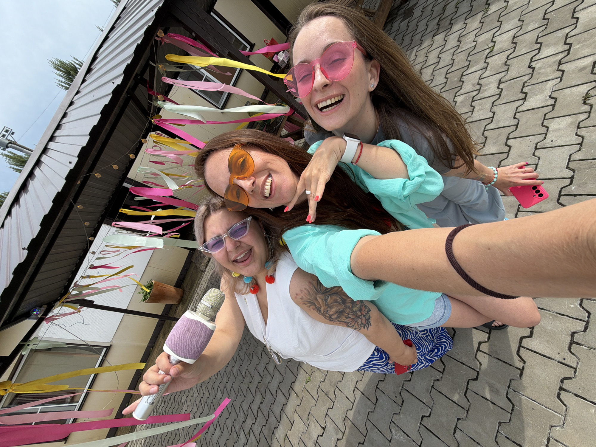 Three young women, appearing to be in their late twenties or early thirties, are posing for a selfie outdoors. They are standing on a paved area with interlocking gray pavers. Above them, a dark-colored, corrugated metal roof is visible, supported by beams. Colorful streamers in shades of pink, yellow, and light green hang from the roofline, creating a festive atmosphere.
The woman on the left is holding a pink and silver microphone and wearing a white top and blue and white patterned shorts. She has blonde hair and is wearing light purple sunglasses. She has a tattoo of a lion on her left arm.
The woman in the middle has brown hair and is wearing orange, heart-shaped sunglasses. She has on a turquoise top.
The woman on the right has long brown hair and is wearing pink, heart-shaped sunglasses. She is wearing a gray top. She is holding a pink smartphone in her right hand. All three women appear to be laughing or smiling broadly, suggesting a joyful or celebratory occasion. The overall scene suggests an outdoor party or gathering. No specific landmarks or architectural details clearly indicate the city of Birobidzhan, Russia.