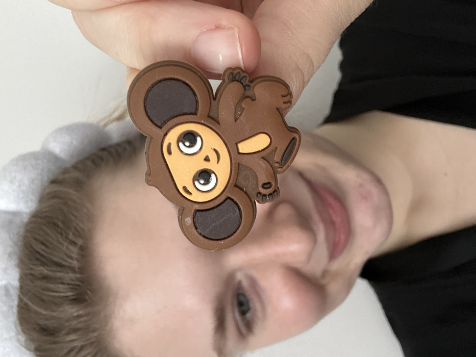 The image displays a close-up, indoor scene focused on a female individual holding a character charm. A fair-skinned female, partially out of focus, occupies the left and lower portions of the frame, smiling with her gaze directed slightly downwards. She has light brown hair and visible blue eyes (specifically the left eye). A textured, light gray headband is worn on her head, and a black garment is visible on her lower body. Centrally positioned and held between the thumb and forefinger of a hand (presumably her right hand) is a brown, rubber or plastic charm depicting the character Cheburashka. The charm features large dark brown ears, an orange-brown face and belly, white eyes with black pupils, and a small black smile. The background is a plain, indistinct light-colored surface, indicative of an indoor setting. There are no visual cues to identify a specific location within the city of Kazan, Russia.
