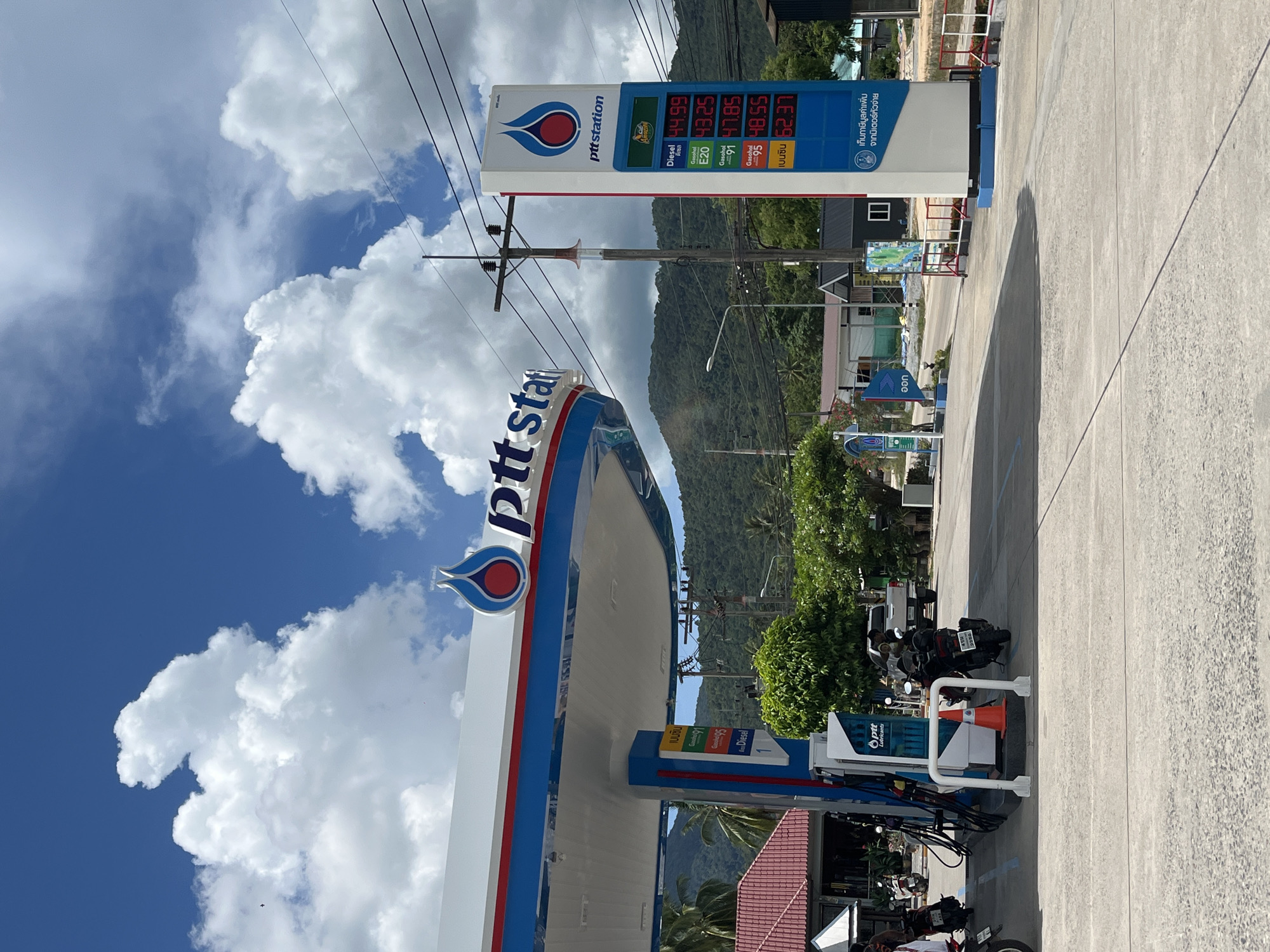 This image depicts a PTT Station gas station in Tambon Ko Phangan, Thailand, under a partly cloudy sky with a bright blue background. The PTT Station signage is prominently displayed on the canopy and a tall price board. The price board shows current fuel prices: Diesel at 44.99, E20 at 43.25, Gasohol 91 at 47.85, Gasohol 95 at 48.55, and a product labeled "เบนซิน" (which translates to "gasoline") at 52.37. Several motorcycles are parked at the station, some with riders present. A white pickup truck is also visible. The environment includes a paved area, green trees, and a mountain range in the background. Power lines and a utility pole are also part of the scene.