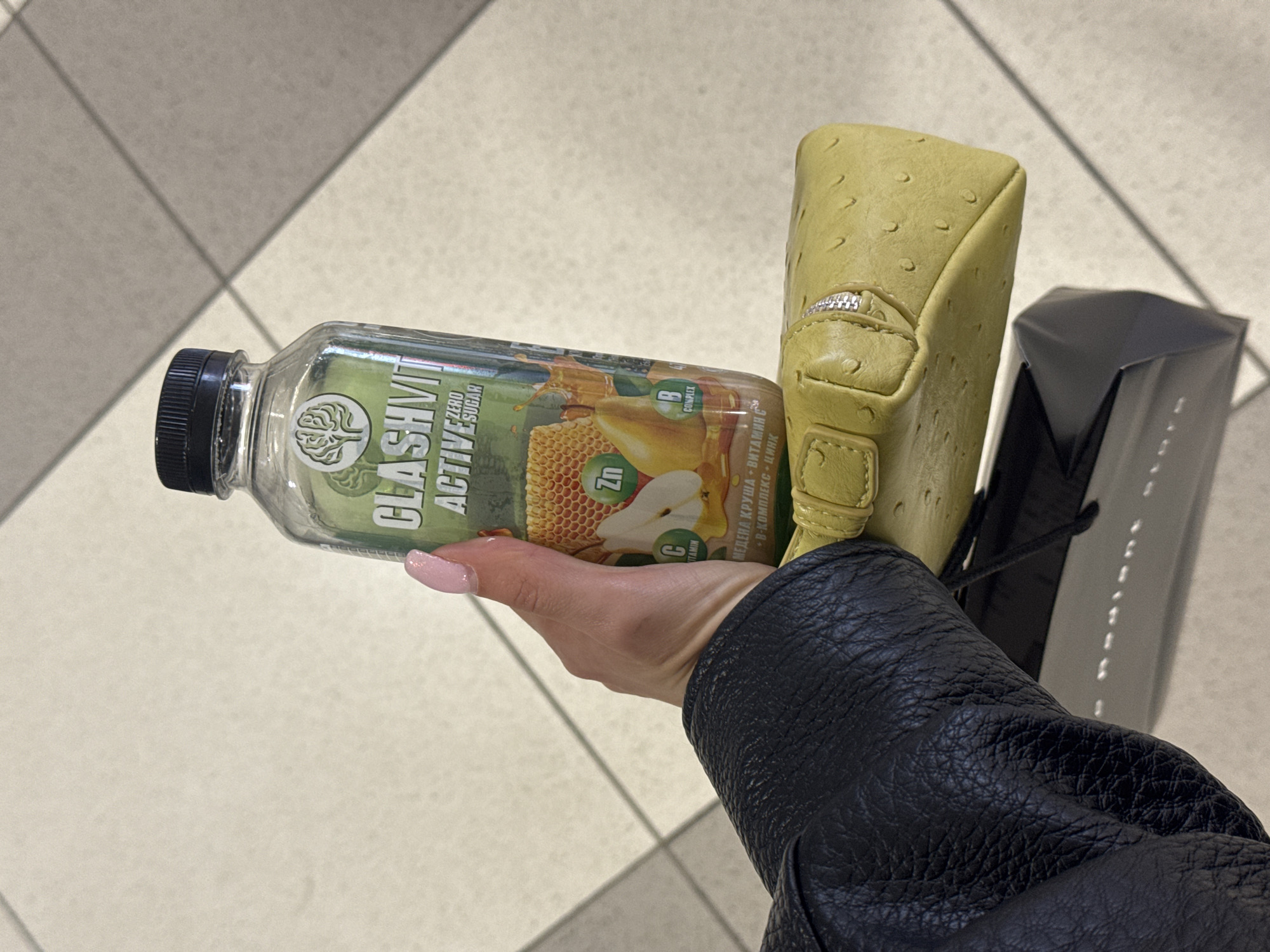 The image captures a person's arm and hand holding several items while likely walking or standing on a tiled floor indoors. The central subject is a clear plastic bottle with a black screw-on cap. Its label, predominantly green, features the brand "GLASMVIT ACTIVE" and "ZERO SUGAR." It also displays icons for "Zn," "B COMPLEX," and "C VITAMIN," alongside images of honeycomb and a pear. Bulgarian text on the label reads "МЕДЕНА КРУША - ВИТАМИН С + В-КОМПЛЕКС + ЦИНК" (Honey Pear - Vitamin C + B-Complex + Zinc).
The hand, adorned with light pink nail polish, also grasps a small, textured, light yellow or lime green pouch or wallet with a visible silver zipper. Partially visible next to the pouch is a dark grey or black shopping bag or box with a black handle. A black, possibly leather, jacket sleeve covers the person's forearm. The setting is an indoor public space, indicated by the light-colored square tiled floor with dark grout lines. The overall scene suggests a casual moment during a shopping trip or outing in an urban environment, aligning with a location like Varna, Bulgaria.