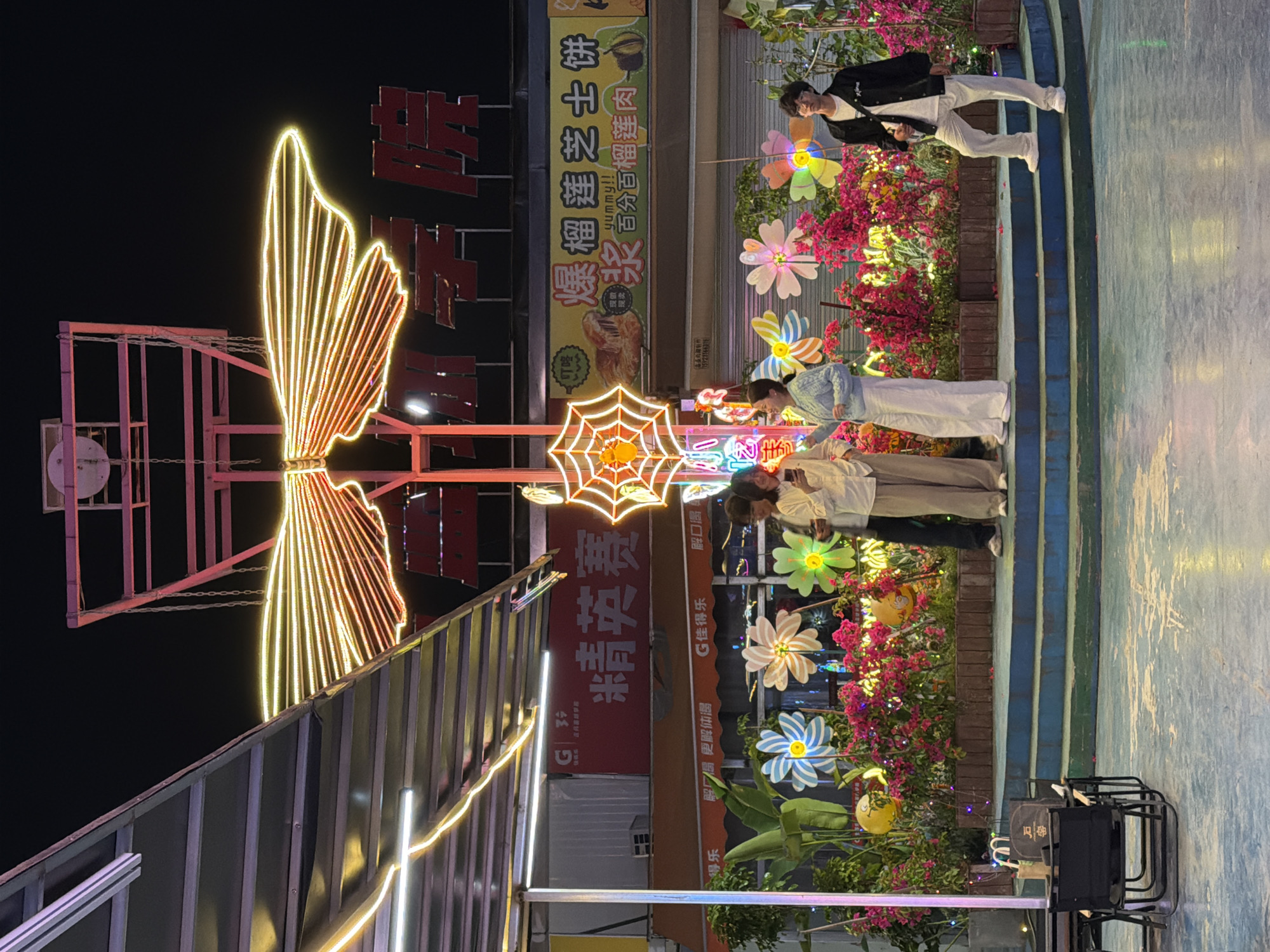 The image depicts an outdoor scene in the city of Chongqing High-tech Zone, China, at night. A group of four people is visible, likely posing for a photograph near a brightly lit decorative area. The background includes various commercial signs in Chinese, colorful flower decorations, and illuminated art installations such as a butterfly and a spider web. A sound system is visible on the bottom right. The ground is a combination of concrete and steps painted with blue and white stripes.