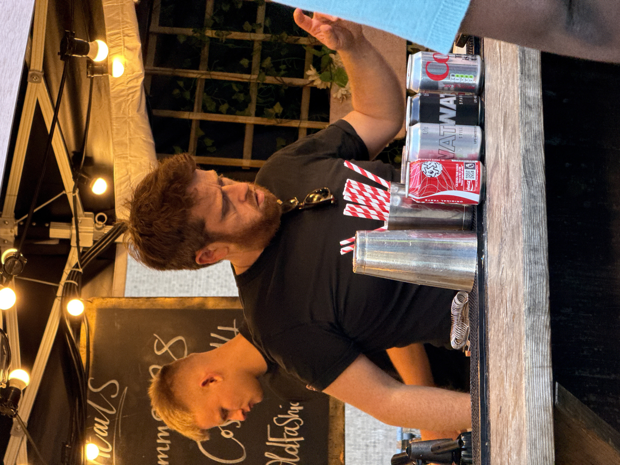 The image shows a man with brown hair and a beard wearing a black t-shirt. He is standing behind a bar, with a Coca-Cola can, Vatna water cans, red and white striped straws, a shaker, and sunglasses on the countertop in front of him. There is also a chalkboard menu. The location appears to be outdoors under a tent with strung lights. A second man is partially visible in the reflection of the chalkboard.