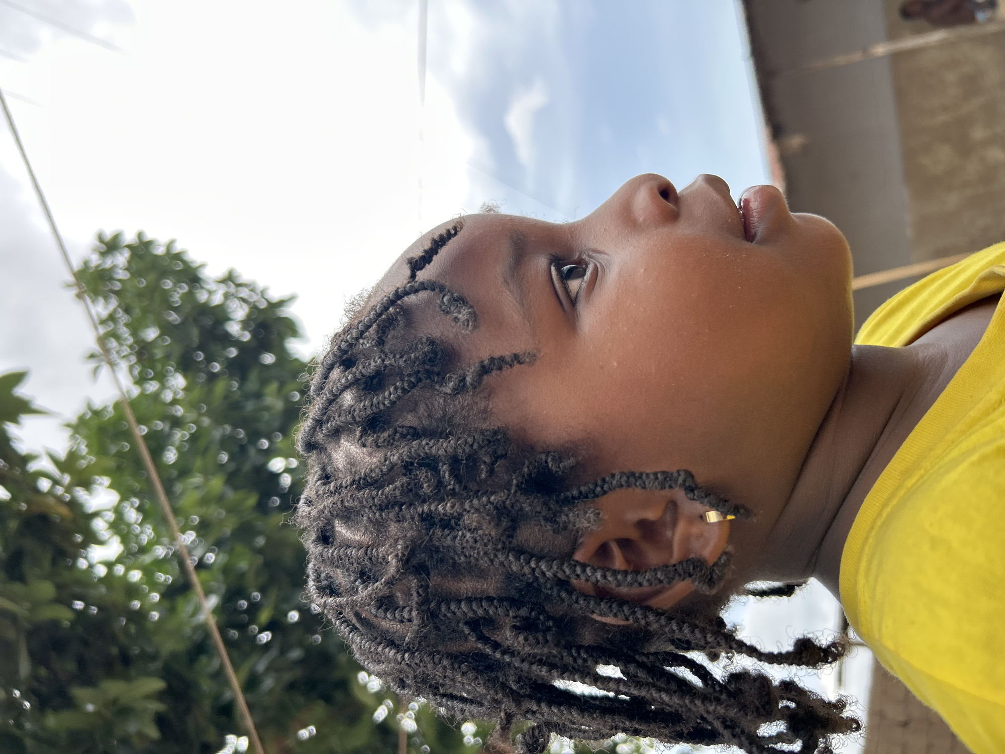 A low-angle, eye-level shot shows a young Black child in Jos, Nigeria. The child's face is oriented upward, eyes gazing towards the sky. The child has dark hair braided close to the scalp and is wearing a yellow garment. To the left, out-of-focus green foliage is visible. Above the child is a patch of sky with light clouds. A blurred building edge appears in the upper right corner.