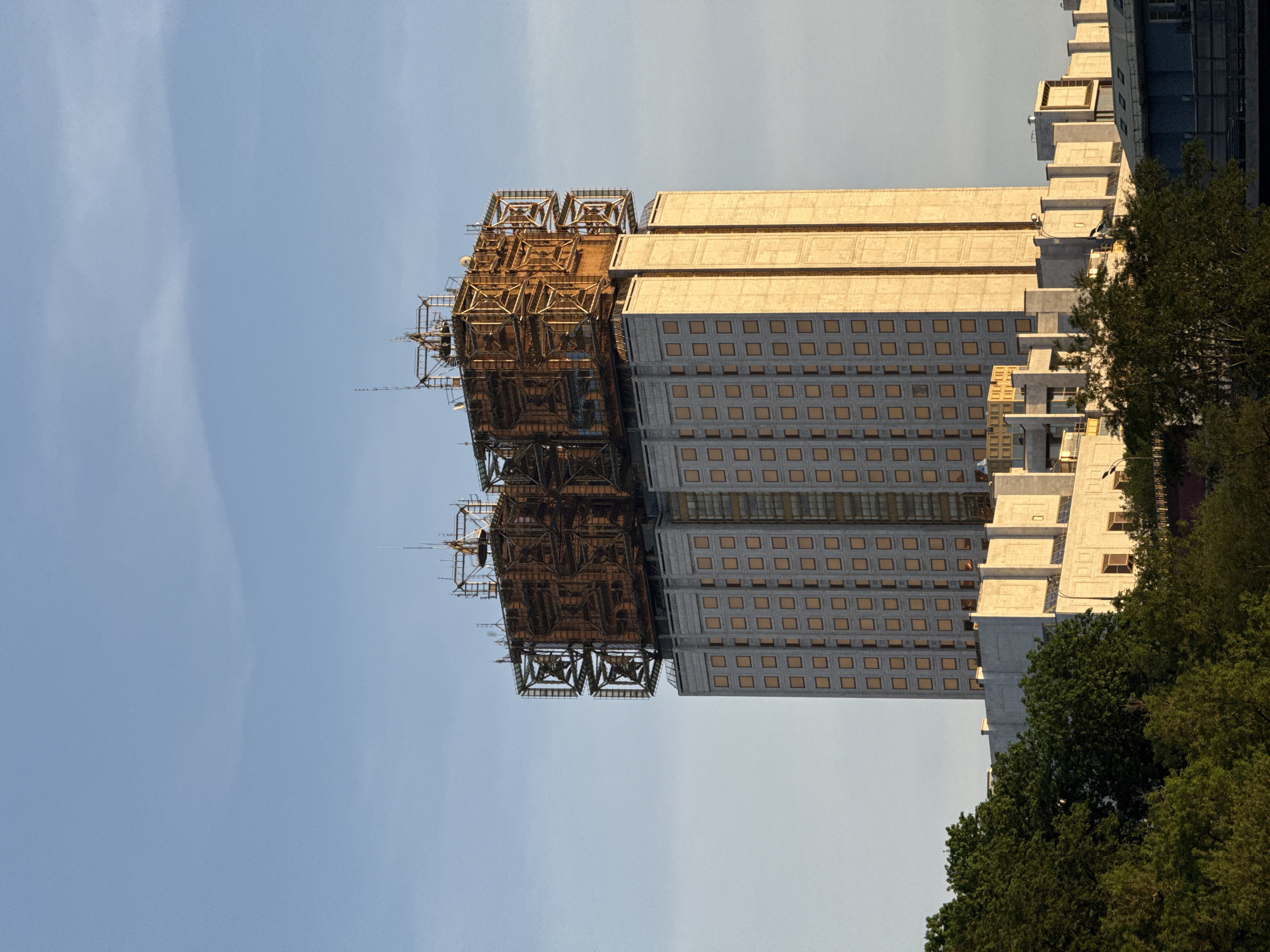 The image depicts the Russian Academy of Sciences building in Moscow, Russia. The architecture showcases a high-rise structure topped with an elaborate, cage-like framework. The main building consists of multiple rectangular tiers, each containing a grid pattern of numerous windows. The cage-like structures atop the building exhibit an intricate network of metal supports.
The sky is a pale blue, suggesting either early morning or late afternoon. The building's facade reflects the warm light of the sun. Patches of green foliage, likely from nearby trees, are visible at the base of the structure.