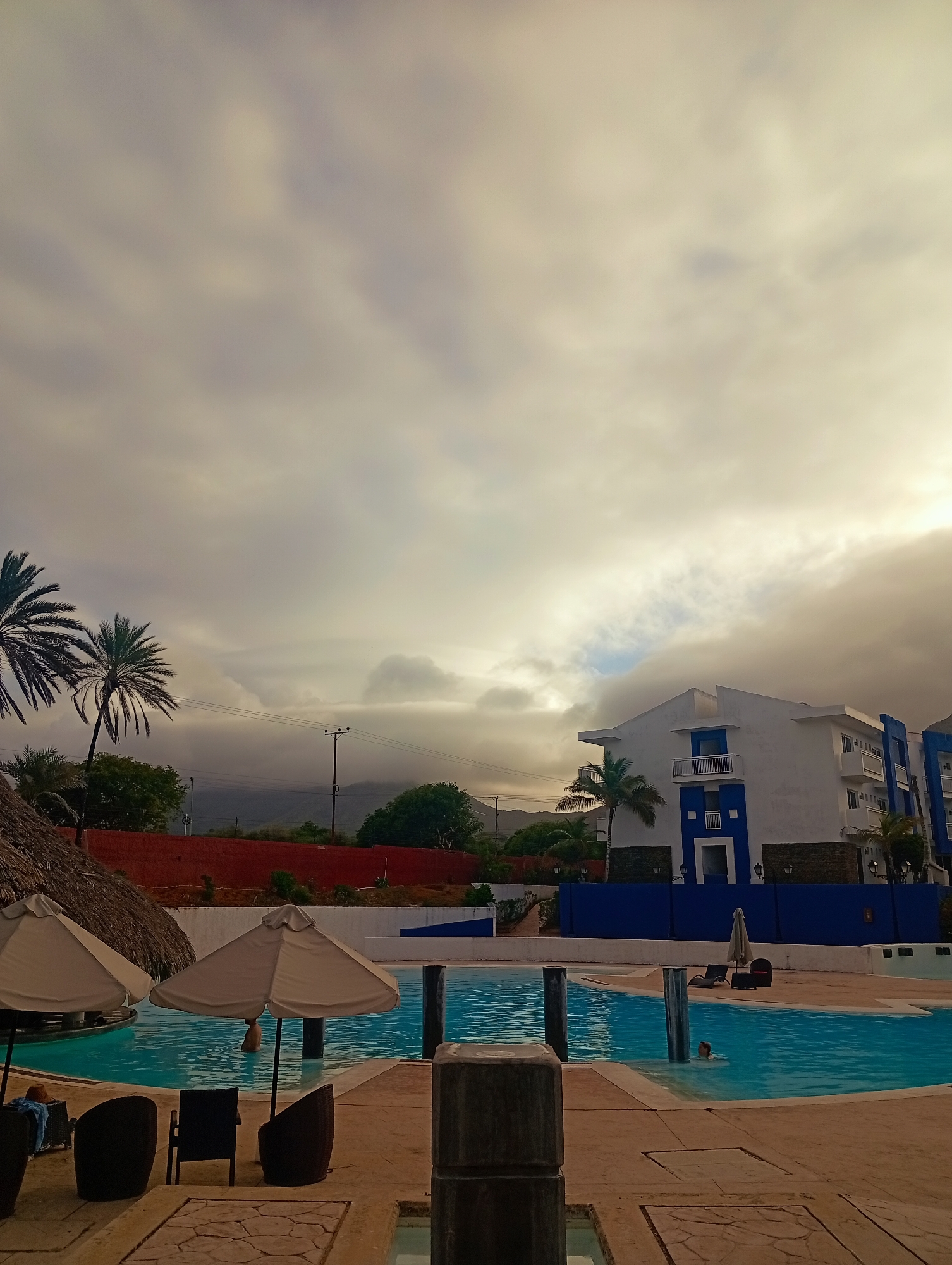 The image depicts a swimming pool area in Playa El Agua, Venezuela, under a cloudy sky. In the foreground, several thatched umbrellas and lounge chairs sit beside a turquoise-colored pool. A person is visible in the water. In the background is a building with white walls and blue trim. Beyond the building, trees and a red wall are visible, with mountains in the distant background. Utility poles are also present. The image suggests a resort or vacation setting.