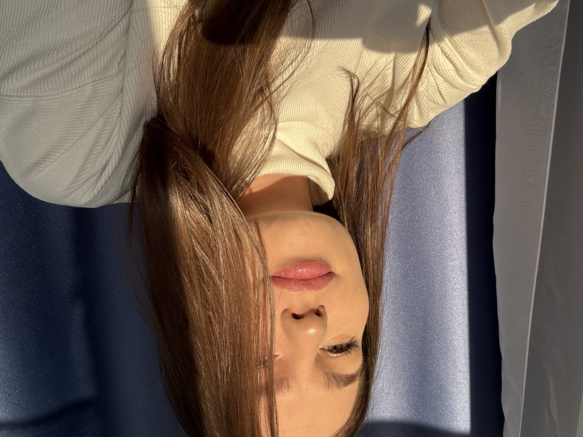 A woman with long brown hair is seen from an inverted perspective. Her face is centered in the frame, with her lips, nose, and one eye visible. She is wearing a white turtleneck sweater. The background consists of a blue surface and a white curtain on the right side of the frame. The location is not identifiable from the image.