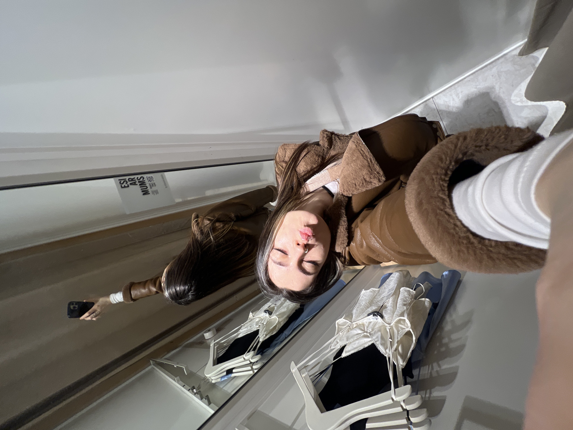 The image depicts a mirror selfie taken inside a retail changing room in Riga, Latvia. A young female subject with long, dark hair, wearing a brown faux-leather jacket with light brown fur trim over a white top, is captured in the reflection. She holds a black smartphone in her extended right hand, angled upwards, taking the photograph. Her eyes are closed, and her lips are pursed in a "kissing" expression, with her head slightly tilted. A silver nose ring is visible on her left nostril.
The environment consists of multiple mirrors reflecting the subject and the interior space. To the left of the primary reflection (on the right side of the actual room), white plastic clothing hangers display various garments, including a black top and a white, lace-trimmed camisole or bralette. The walls appear light-colored, possibly white or off-white. A white rectangular sticker is affixed to a mirrored surface or door frame, displaying the Latvian text "ESI AR MŪMS" (Be With Us), a QR code, and "WWW.MUMS.LV". This URL corresponds to a children's clothing store named "MŪMS" with known locations in Riga, including within Galerija Centrs. However, the clothing visible on the hangers within the fitting room appears to be adult women's fashion. The floor is light-toned tile or laminate.