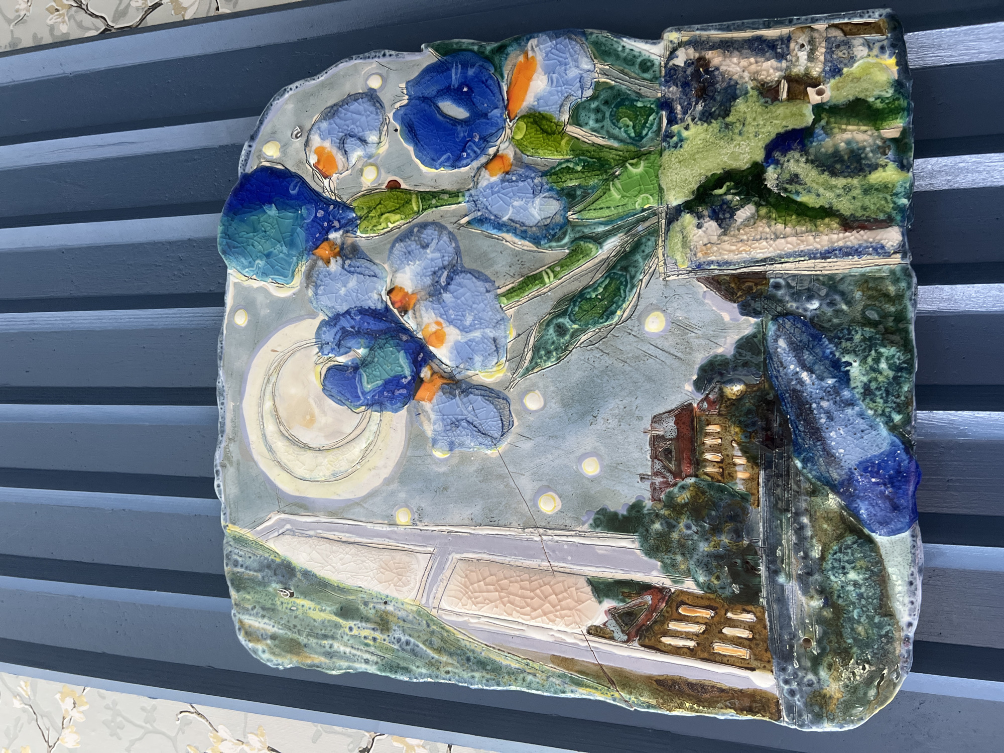 The image shows a ceramic art piece depicting a still life with a nocturnal landscape visible through a window. The ceramic piece features a bouquet of blue irises with orange accents in the foreground, placed in a vase. Behind the flowers, a window reveals a moonlit scene with houses and trees. The overall style is textured and somewhat impressionistic, with a rich use of glazes. The art piece is displayed against a blue, ribbed background, possibly part of a wall or furniture. There is also a hint of wallpaper with a floral pattern visible to the left. There are no people or discernible actions in the image. The ceramic artwork itself is the primary subject, showcasing artistic expression rather than a specific location in Veliky Novgorod, Russia, or any other real-world setting.