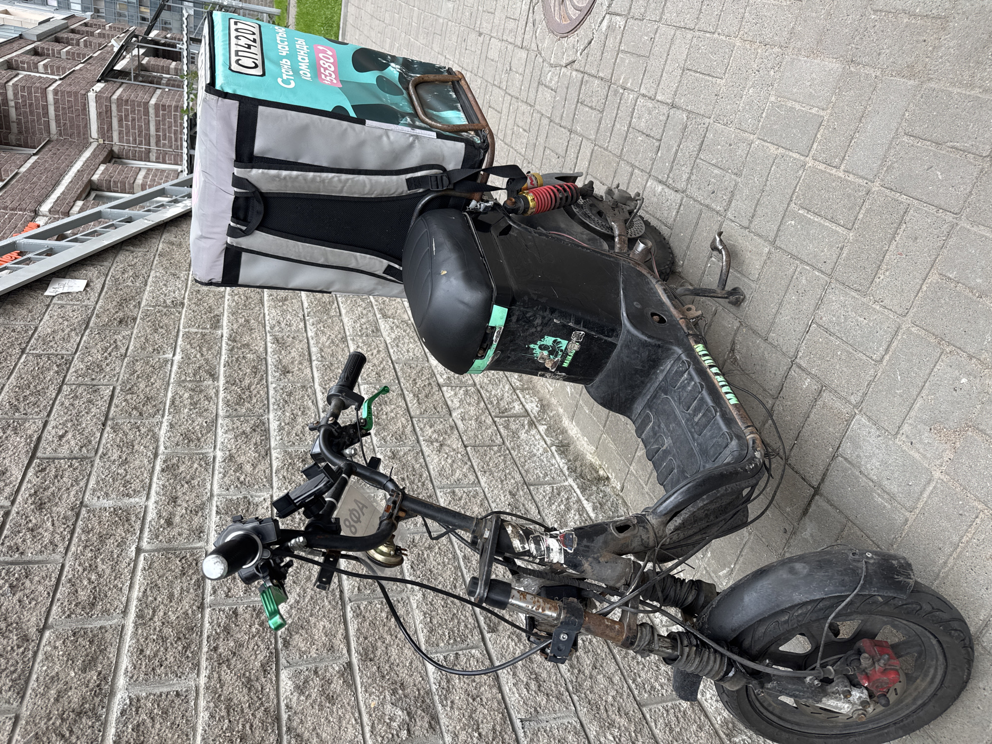 A weathered black electric scooter, equipped with a large, insulated delivery bag, is parked outdoors against a textured brick wall. The scooter exhibits signs of use, including rust and scuffs, particularly on its front forks and frame. The delivery bag, predominantly grey with teal accents, displays text in Russian, including "СП4207" and "Стань частью команды" (Become part of the team), along with a phone number "5580J." The scooter's seat is black, and it has a visible rear suspension. The ground is paved with interlocking grey bricks. In the background, a modern brick building with a metallic staircase attached to its facade is visible. The environment suggests an urban setting, possibly a residential or commercial area.