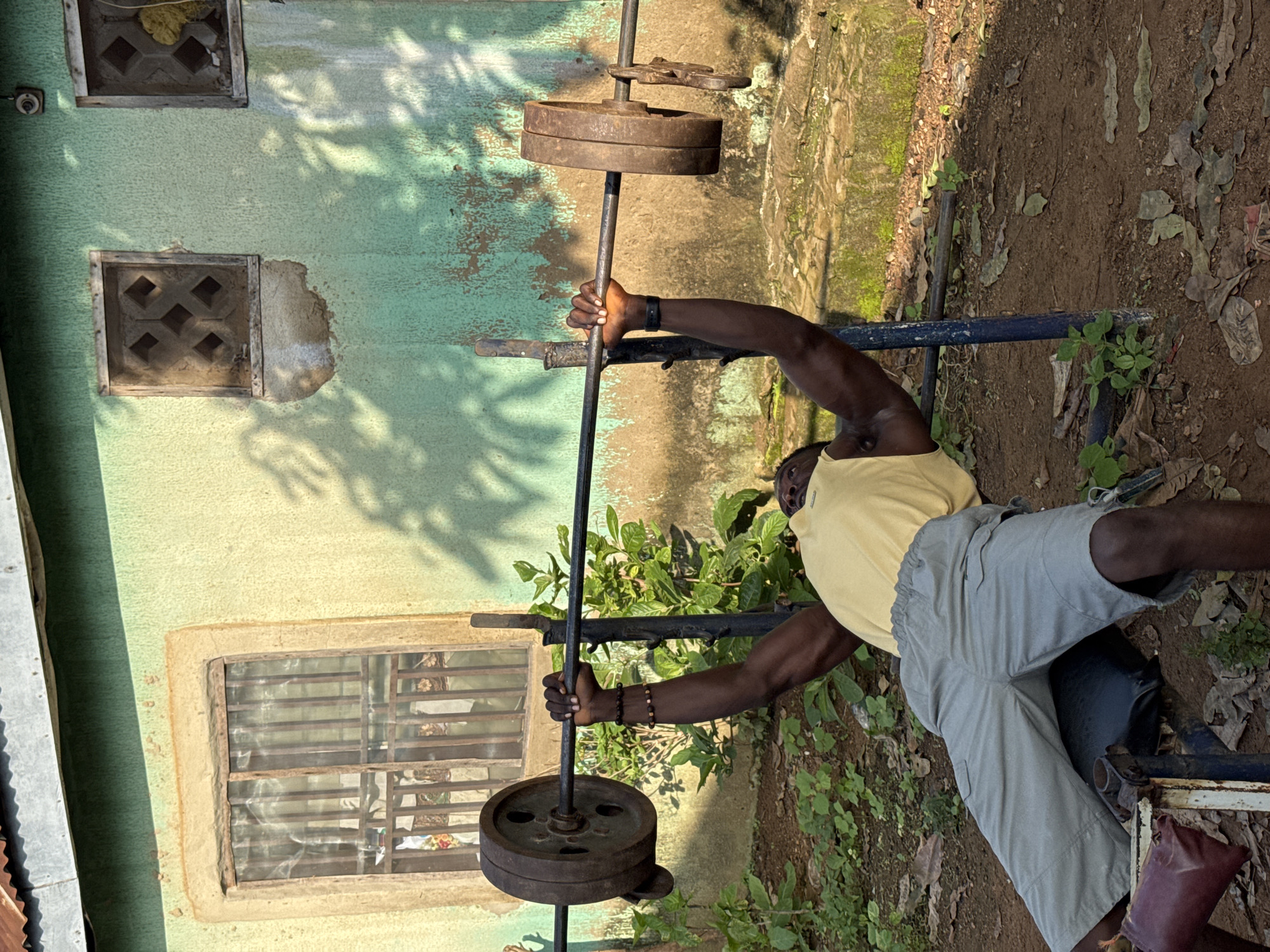 An African man is performing a bench press outdoors. He is lying on a bench press apparatus with a barbell loaded with weight plates across his chest. He is wearing a yellow tank top and light gray cargo shorts. The background shows a weathered, light green building with two small, decorative windows and a larger window with bars and curtains. The ground is covered in dirt and fallen leaves, with some green vegetation visible. The lighting suggests it is daytime, with shadows from leaves cast on the building. This scene is located in the city of Akwanga, Nigeria.